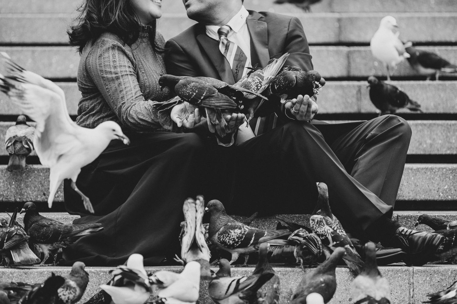 Black and white photo of a couple sitting on steps surrounded by pigeons, with birds perched on their hands.
