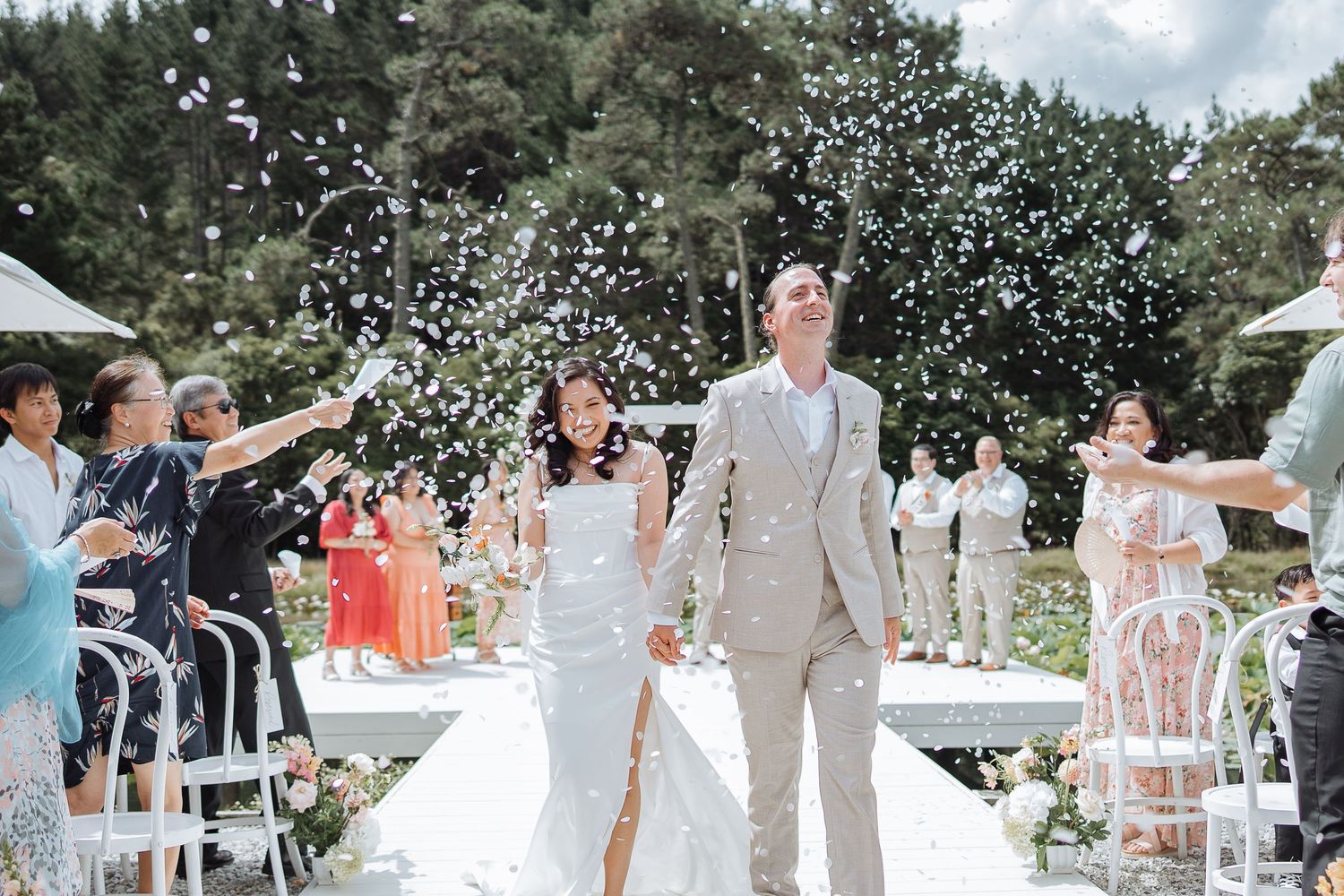 Newlyweds walk down outdoor aisle while guests shower them with confetti at garden wedding ceremony.