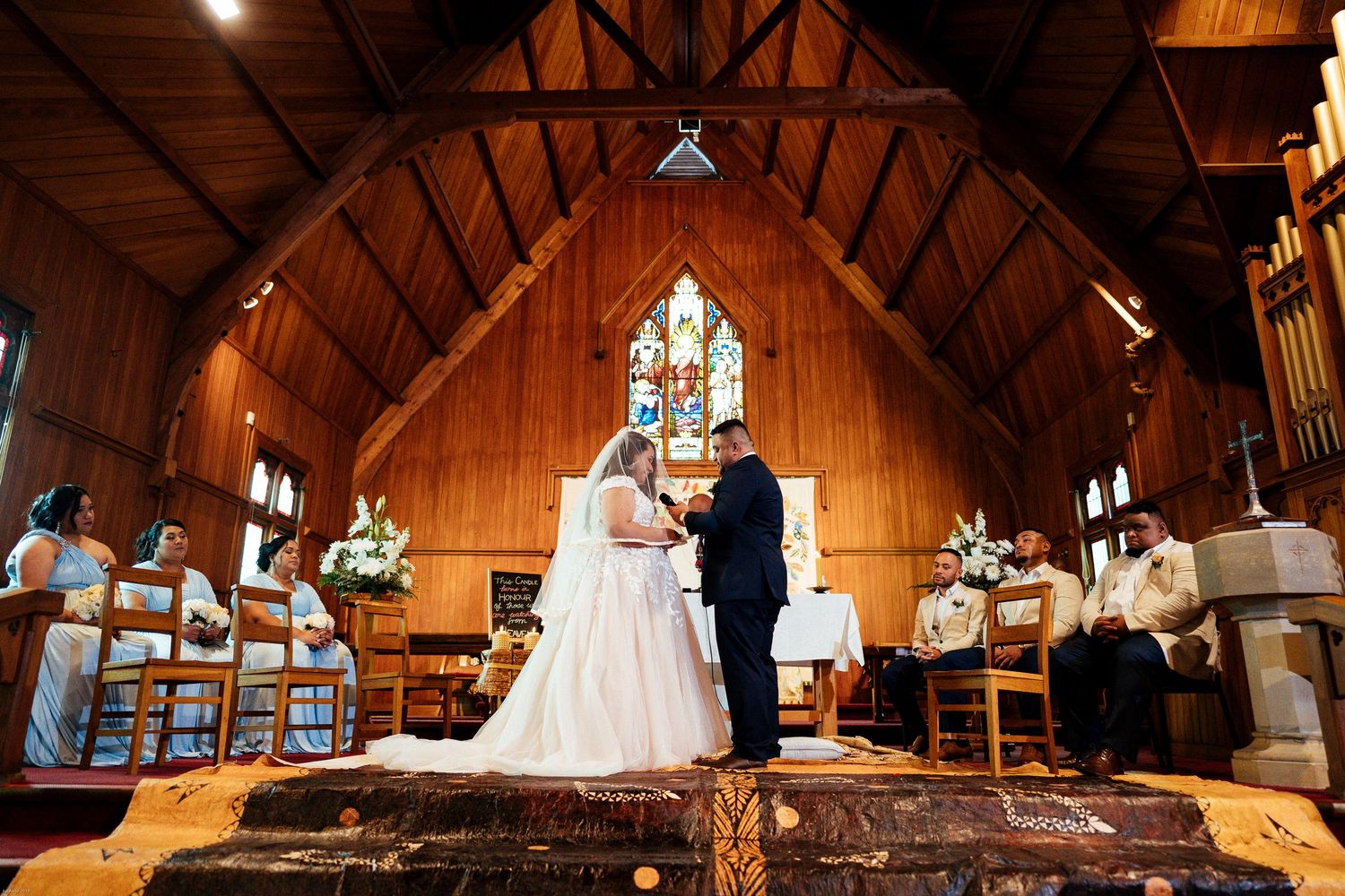 A wedding ceremony takes place in a wooden chapel with stained glass windows and guests seated in wooden chairs.