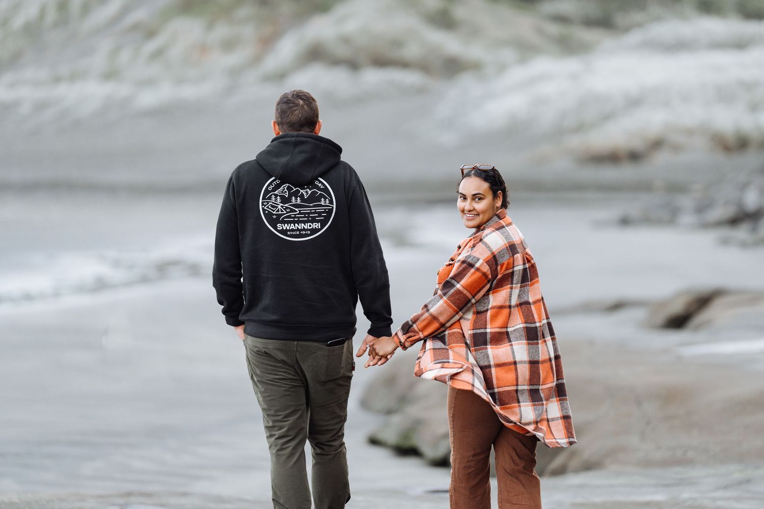 Couple holding hands on a beach, woman in plaid shirt looks back smiling, man in black hoodie walks ahead.