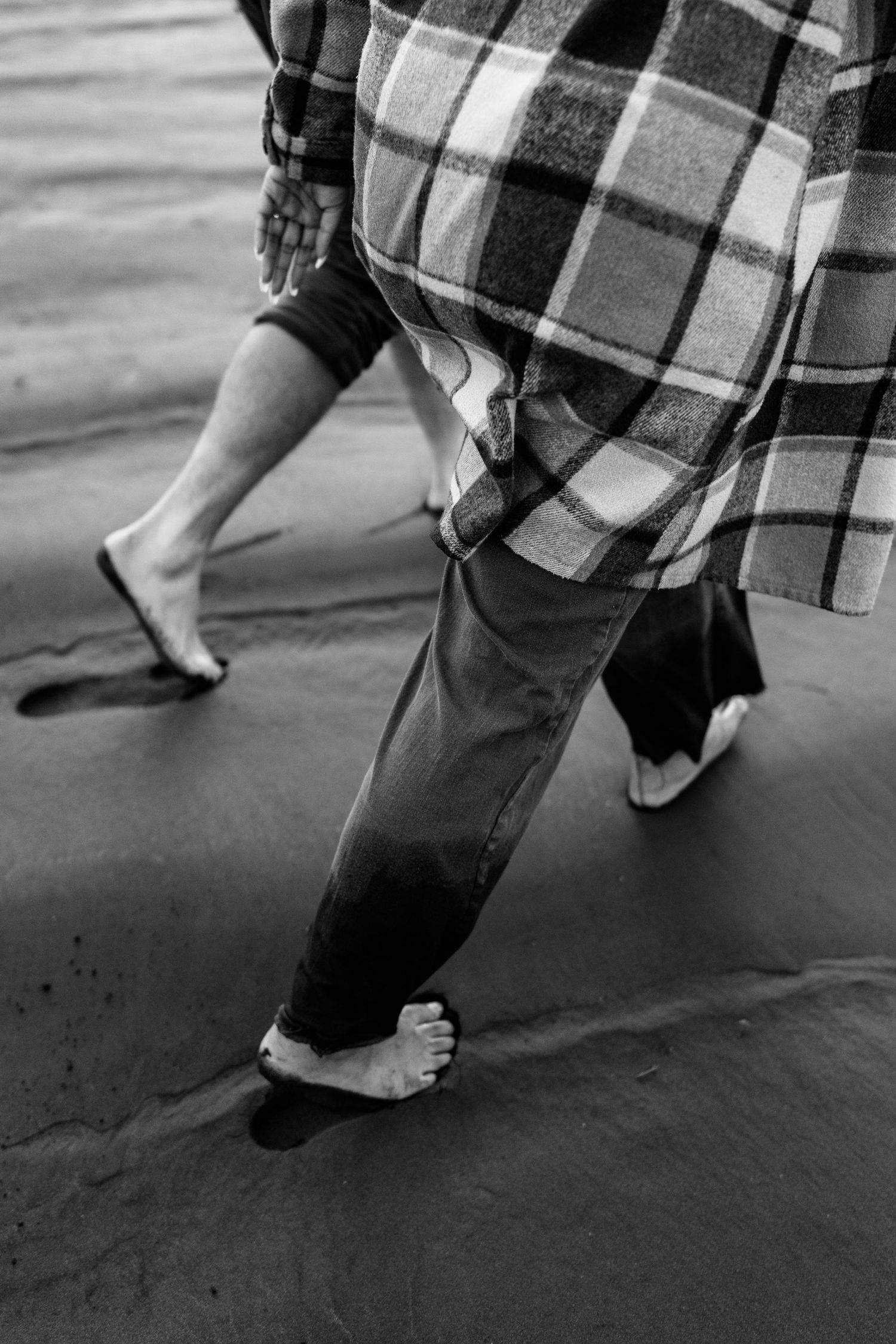 Black and white photo of two people walking barefoot on wet sand beach, one wearing plaid shirt and jeans.