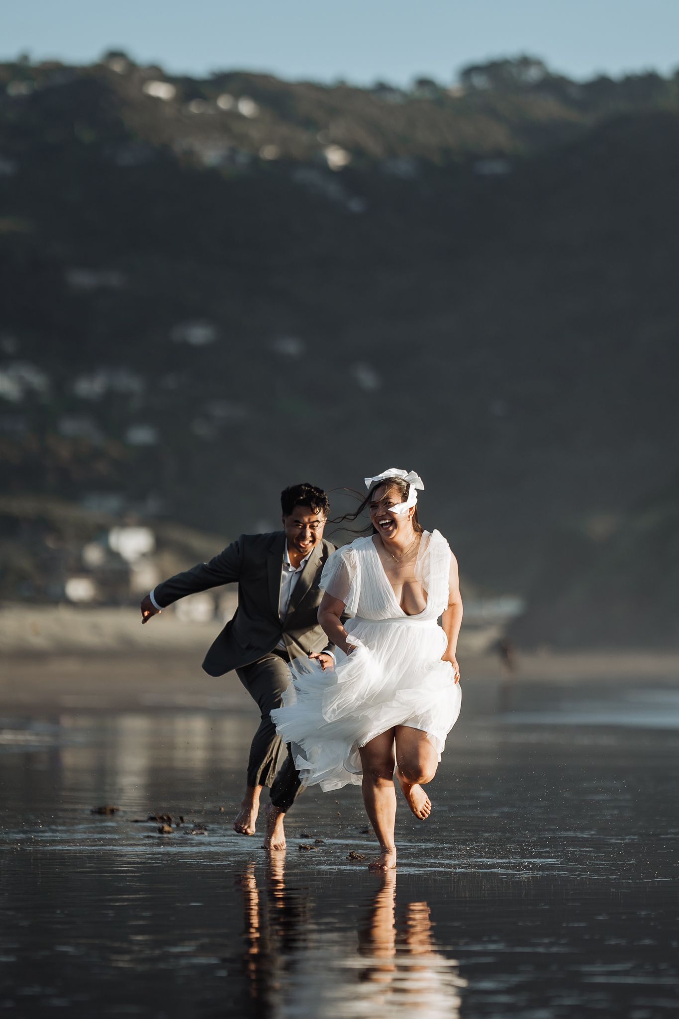 Couple runs playfully through shallow ocean water at beach during sunset photoshoot.