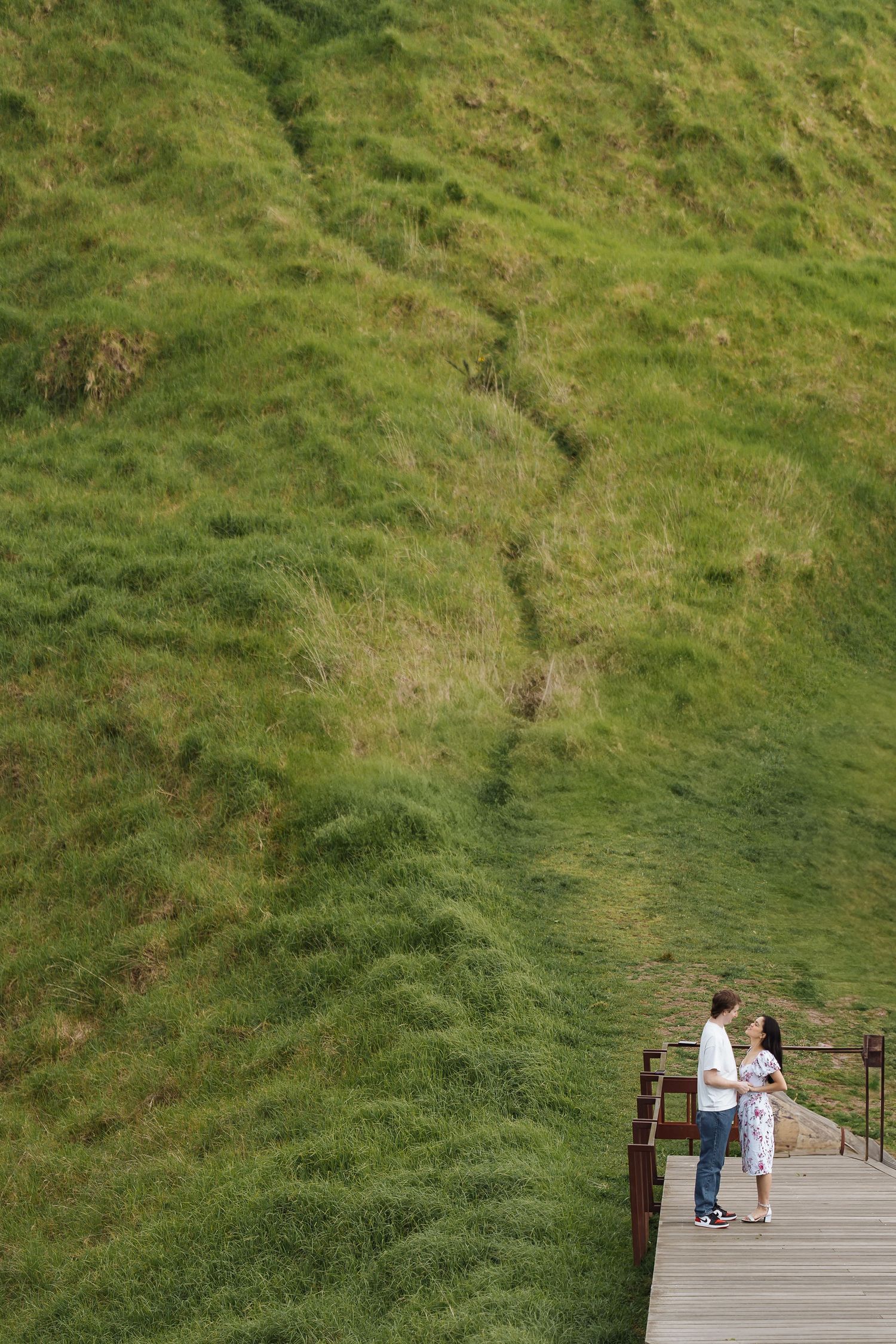 Couple standing on wooden viewing platform surrounded by lush green hillside.