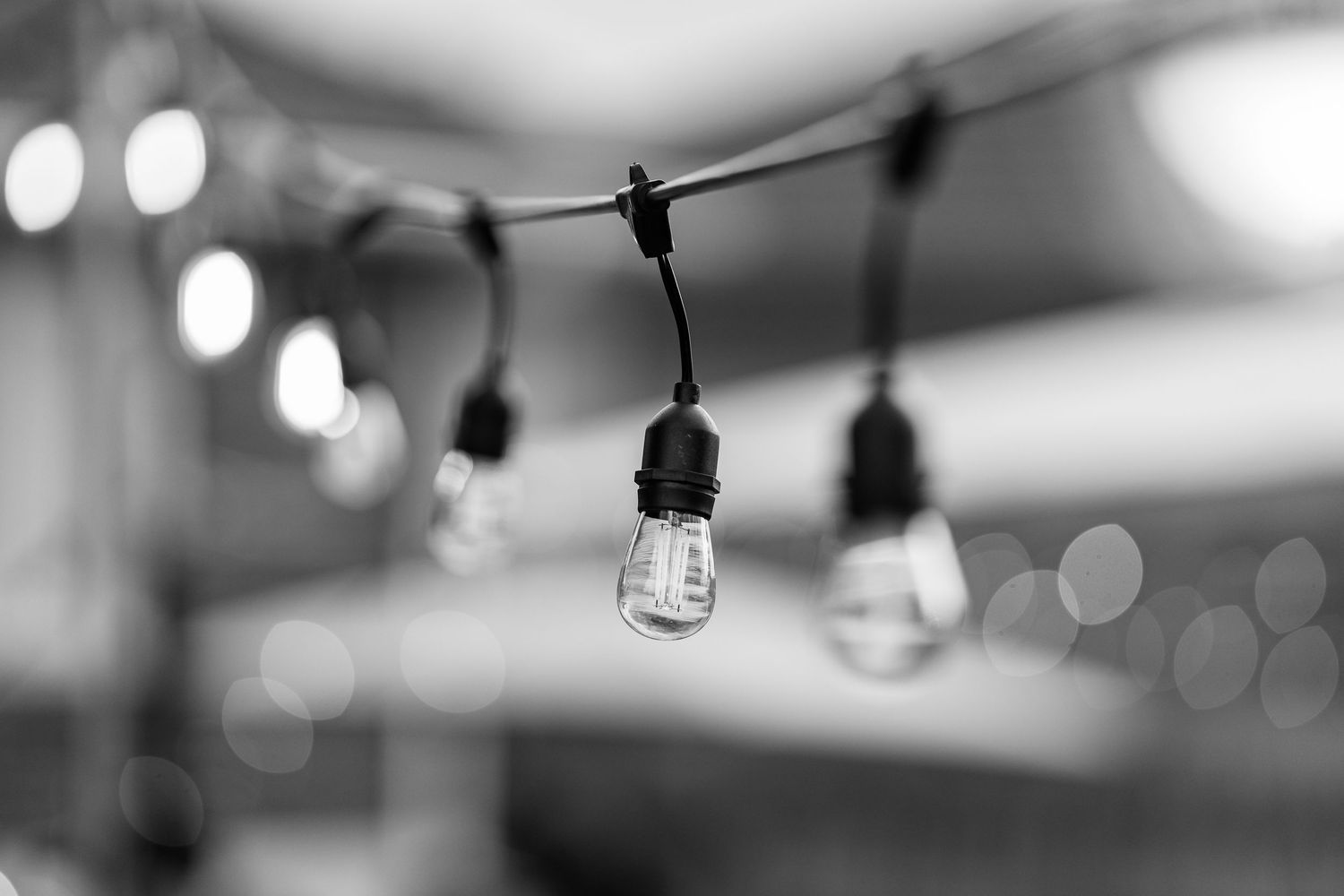 Black and white photo of vintage Edison bulb string lights hanging outdoors with bokeh background.
