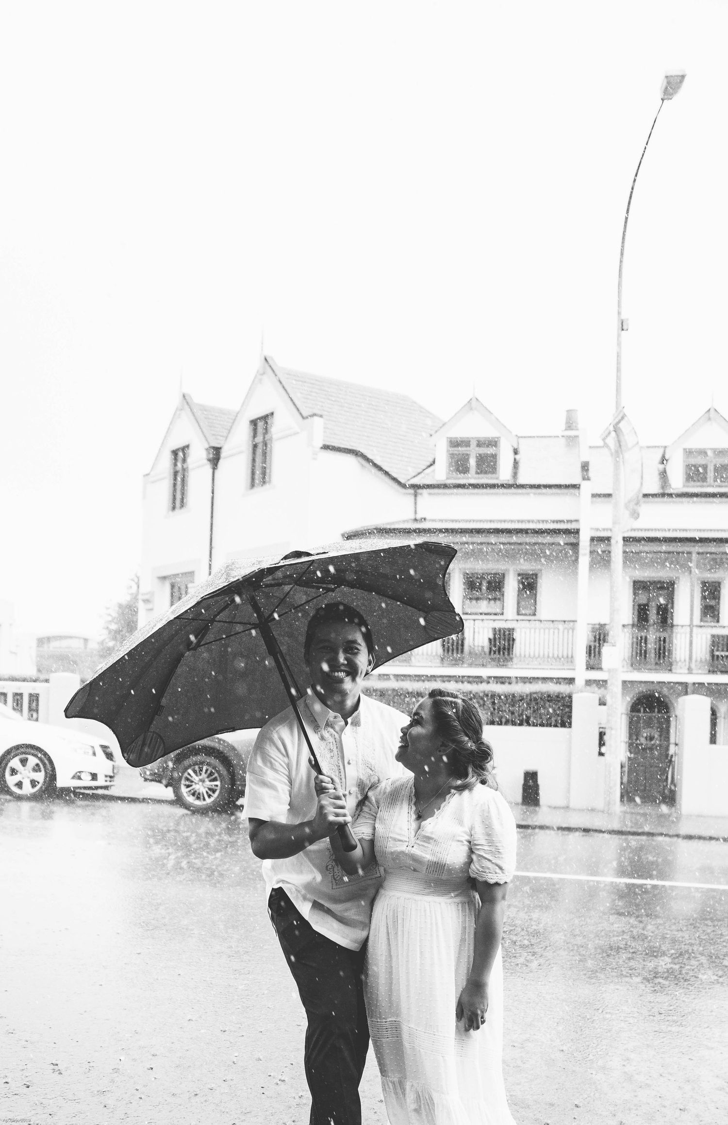 A couple shares an umbrella in the rain outside a white colonial-style building.