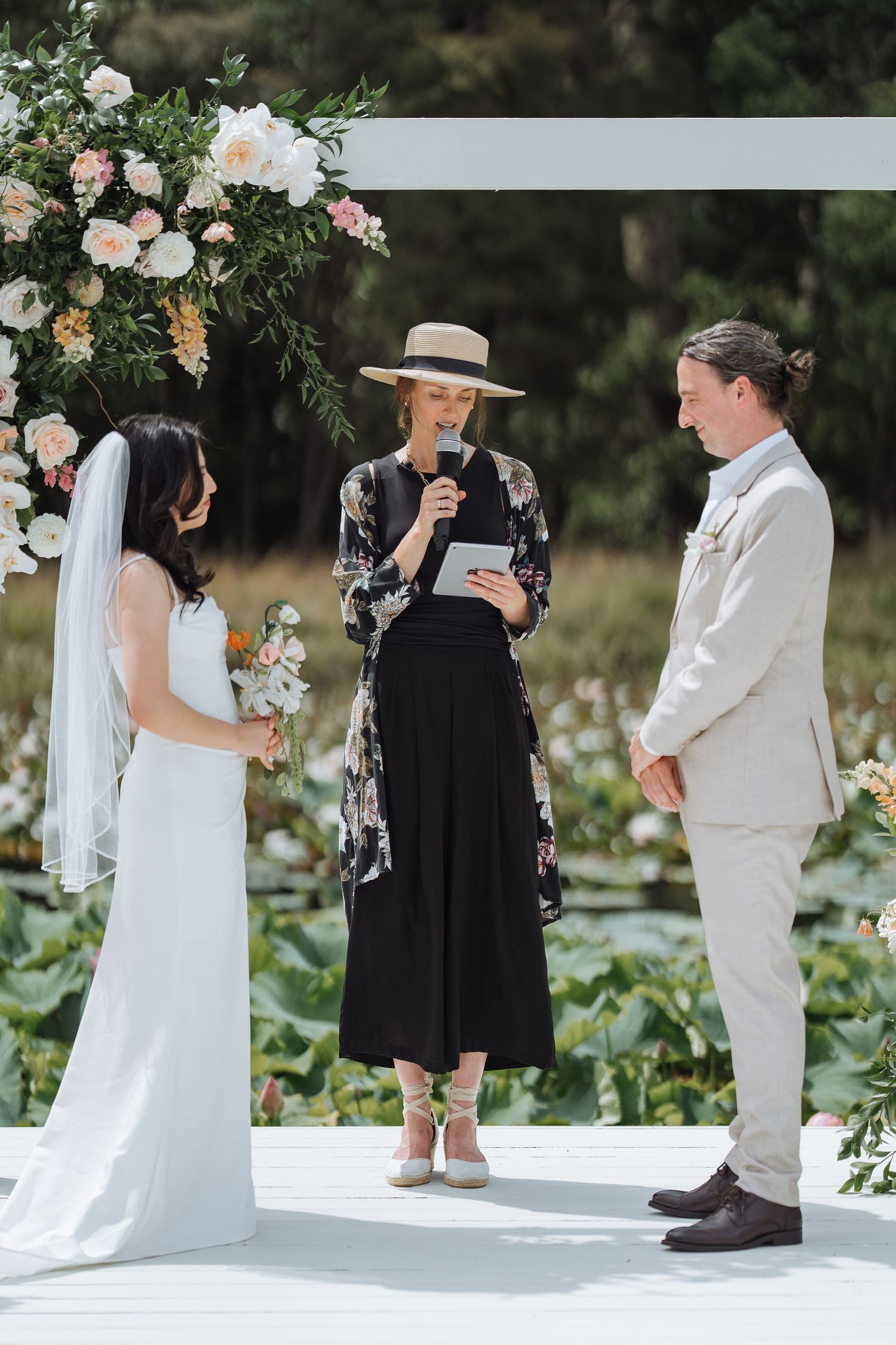 An outdoor wedding ceremony under a floral arch with an officiant in a black dress and wide-brimmed hat.