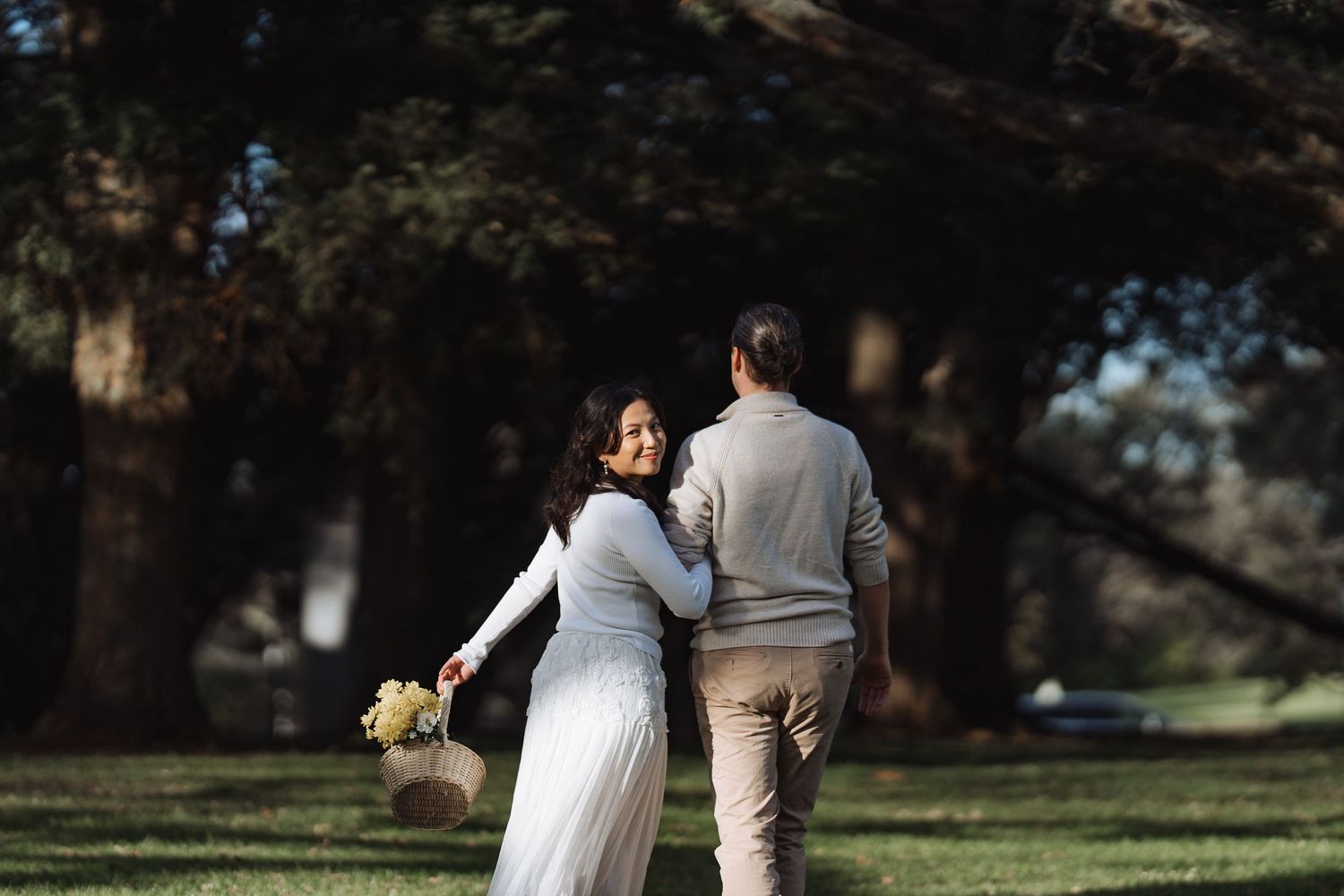 A couple walks hand in hand through a sunlit outdoor garden setting.