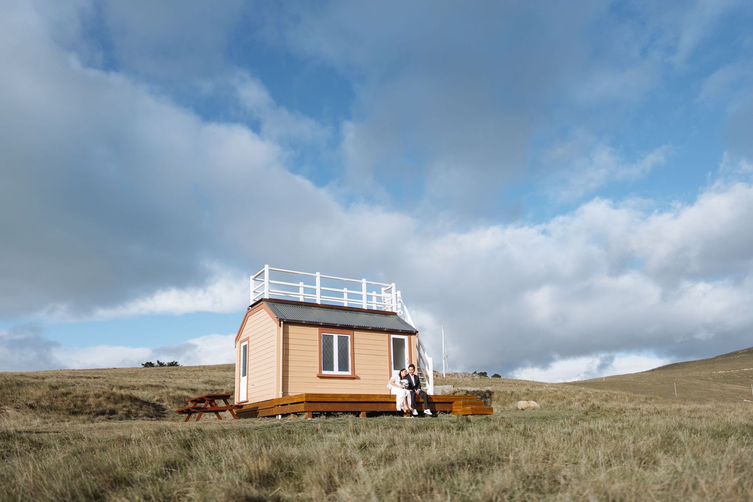 A pink tiny house sits on a grassy hillside under cloudy skies.