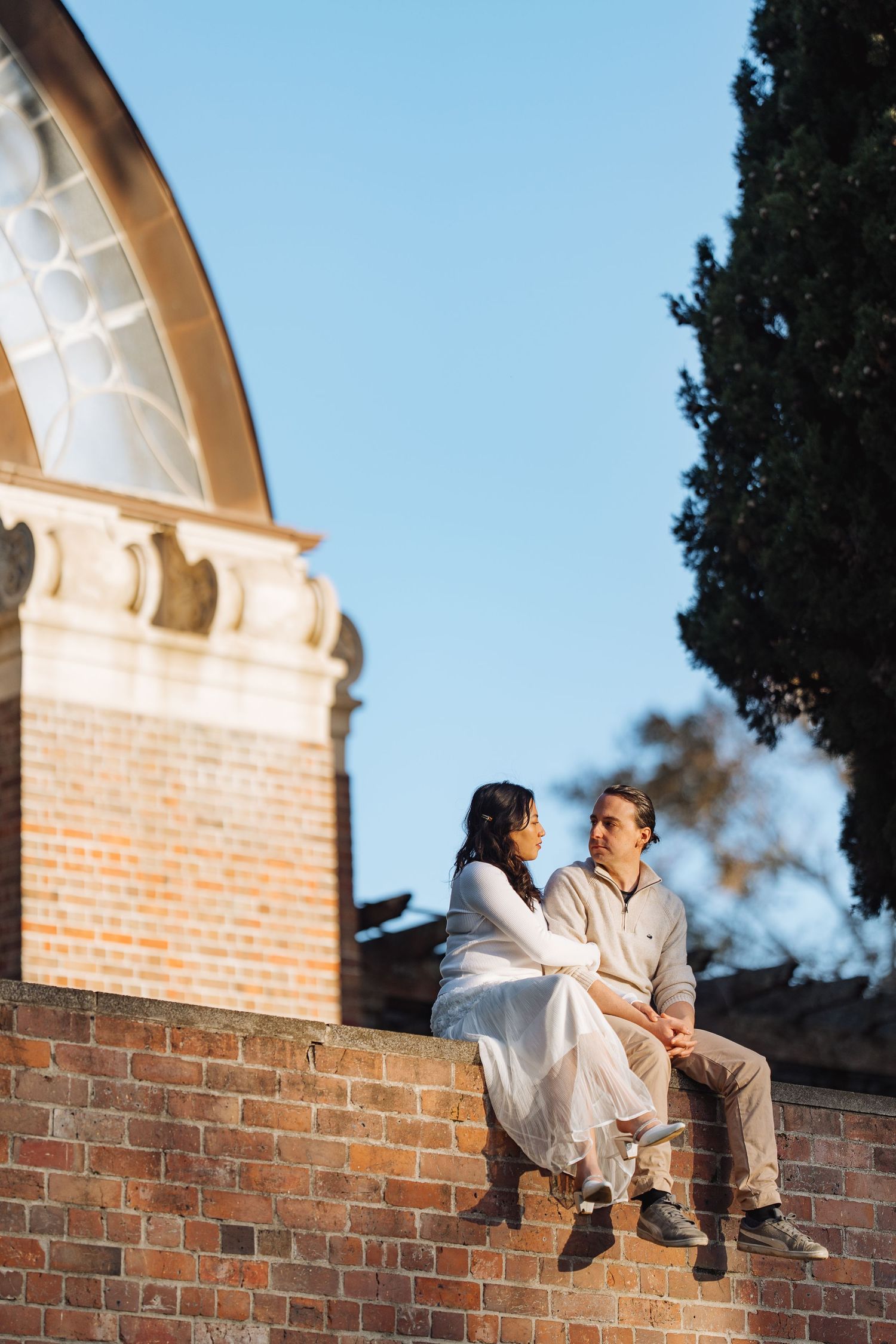 A couple sits atop a brick wall under a blue sky having an intimate conversation.