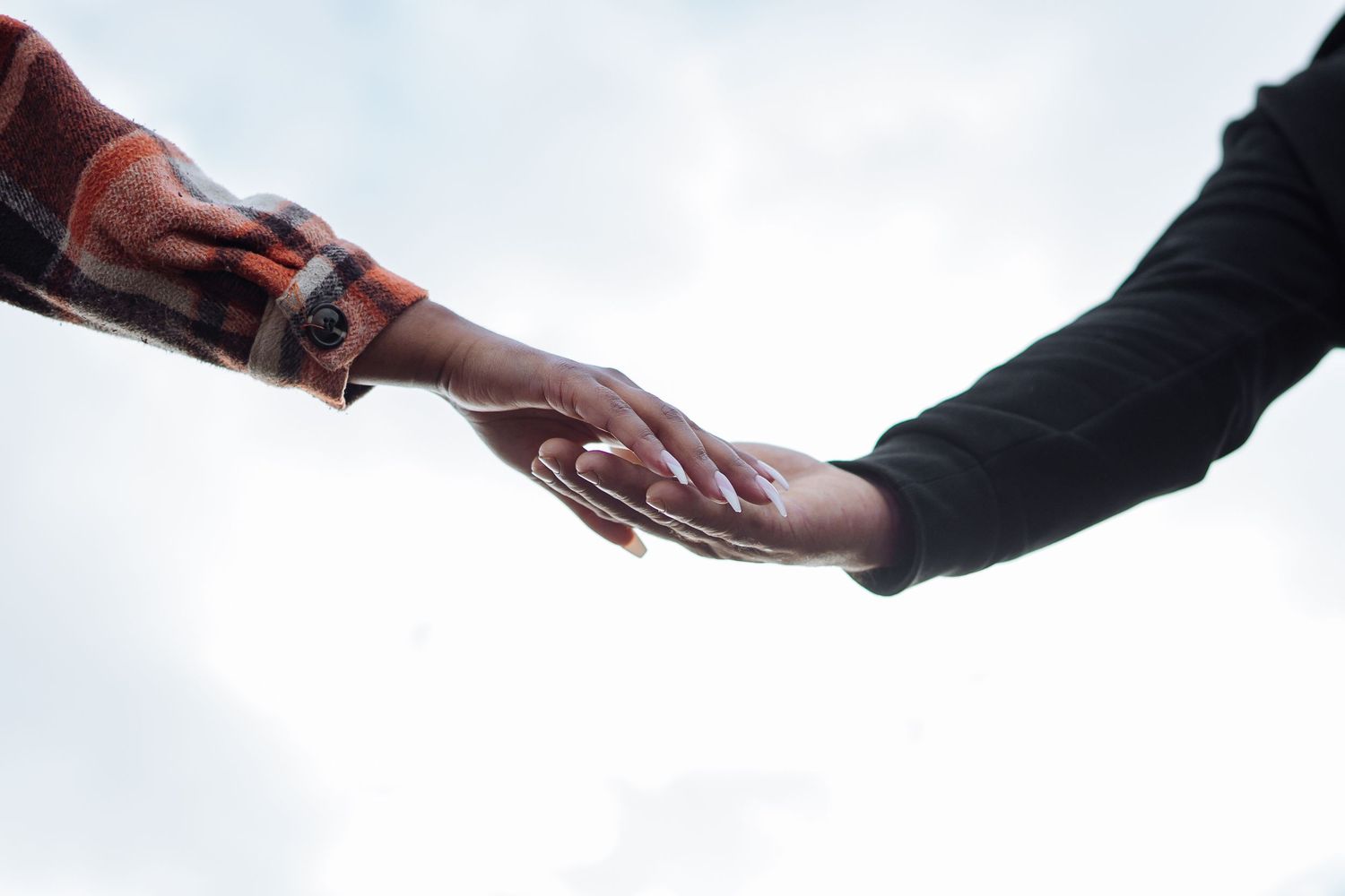 Two people reaching out to touch hands against a cloudy sky, one wearing a red flannel sleeve with tattoos.