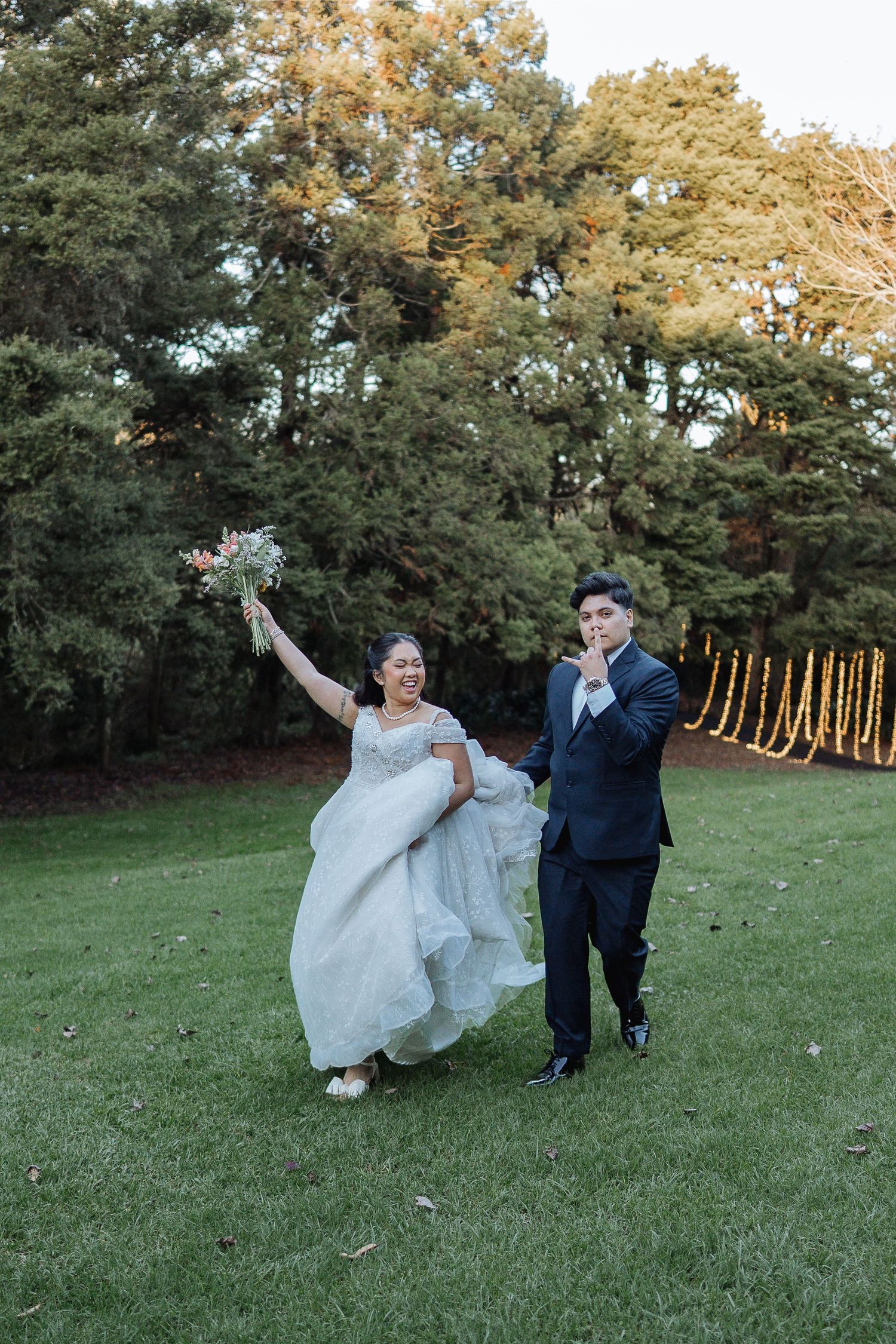 Playful wedding couple dancing and twirling on grass with autumn trees in background.