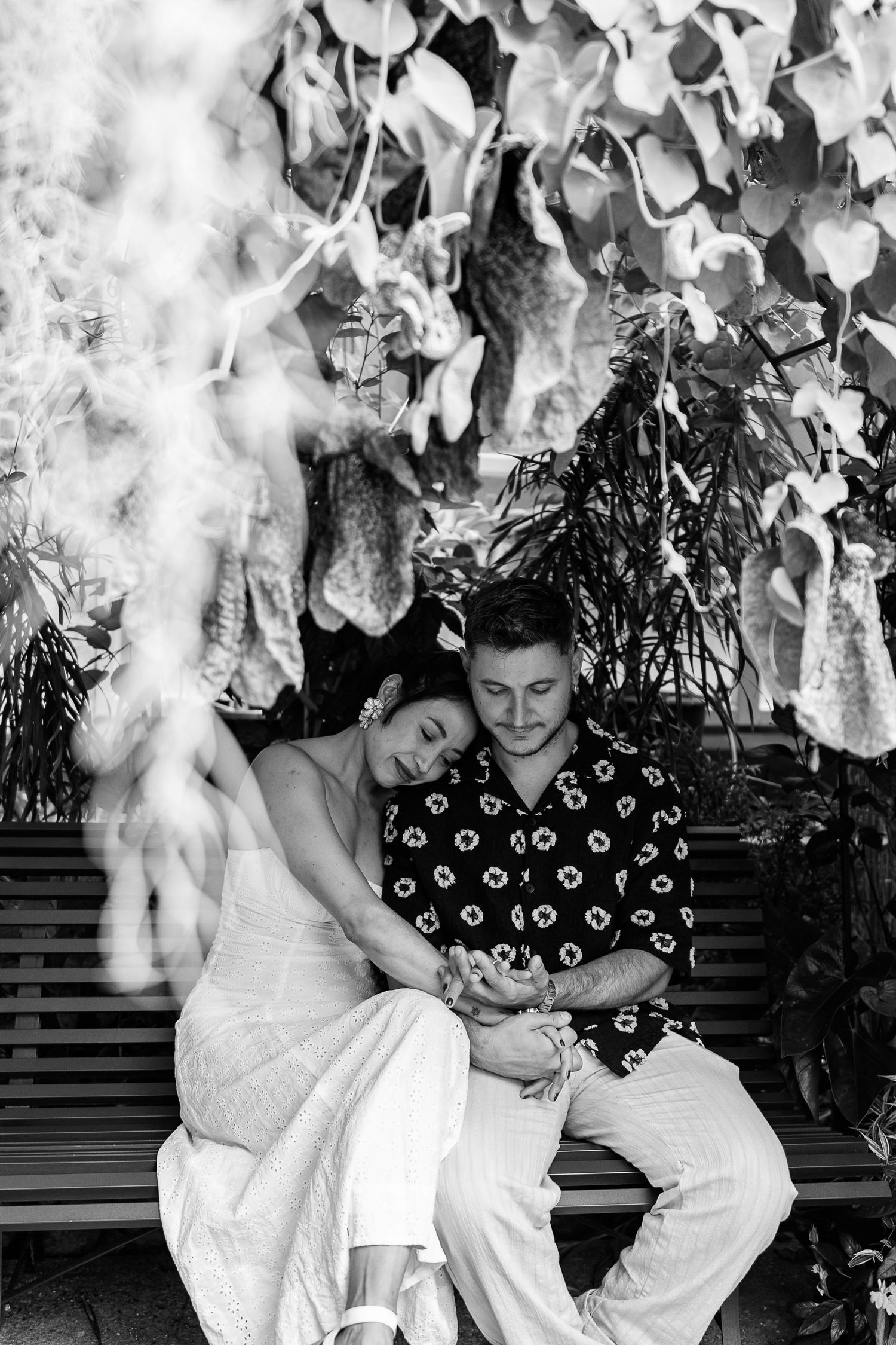 A couple rests on a bench under hanging white flowers in black and white photography.