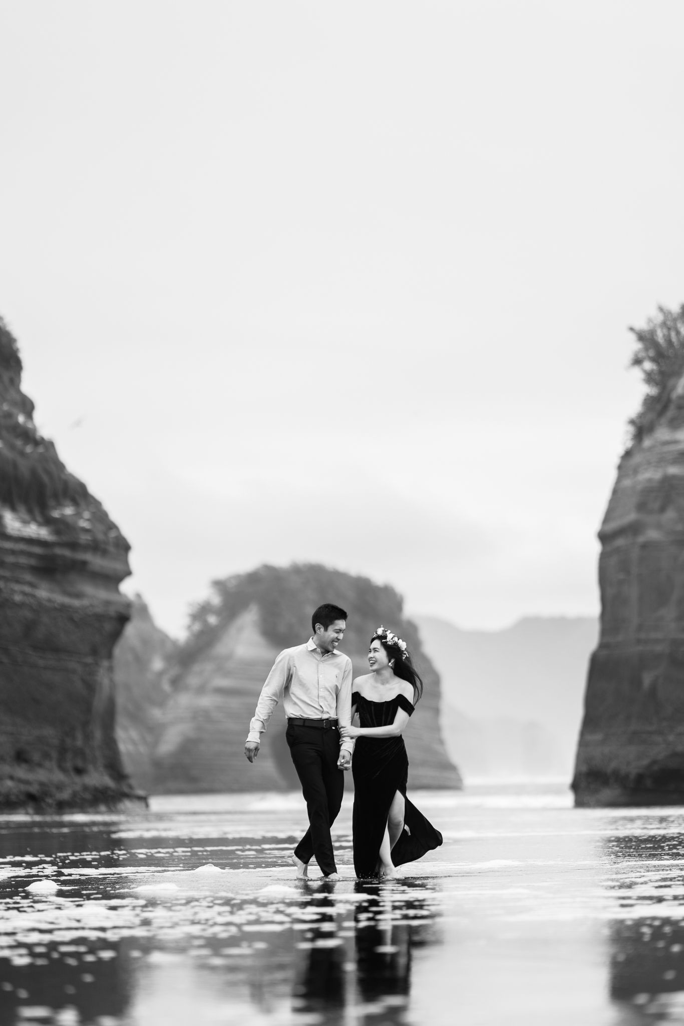 Couple standing in shallow ocean water near dramatic coastal rock formations.
