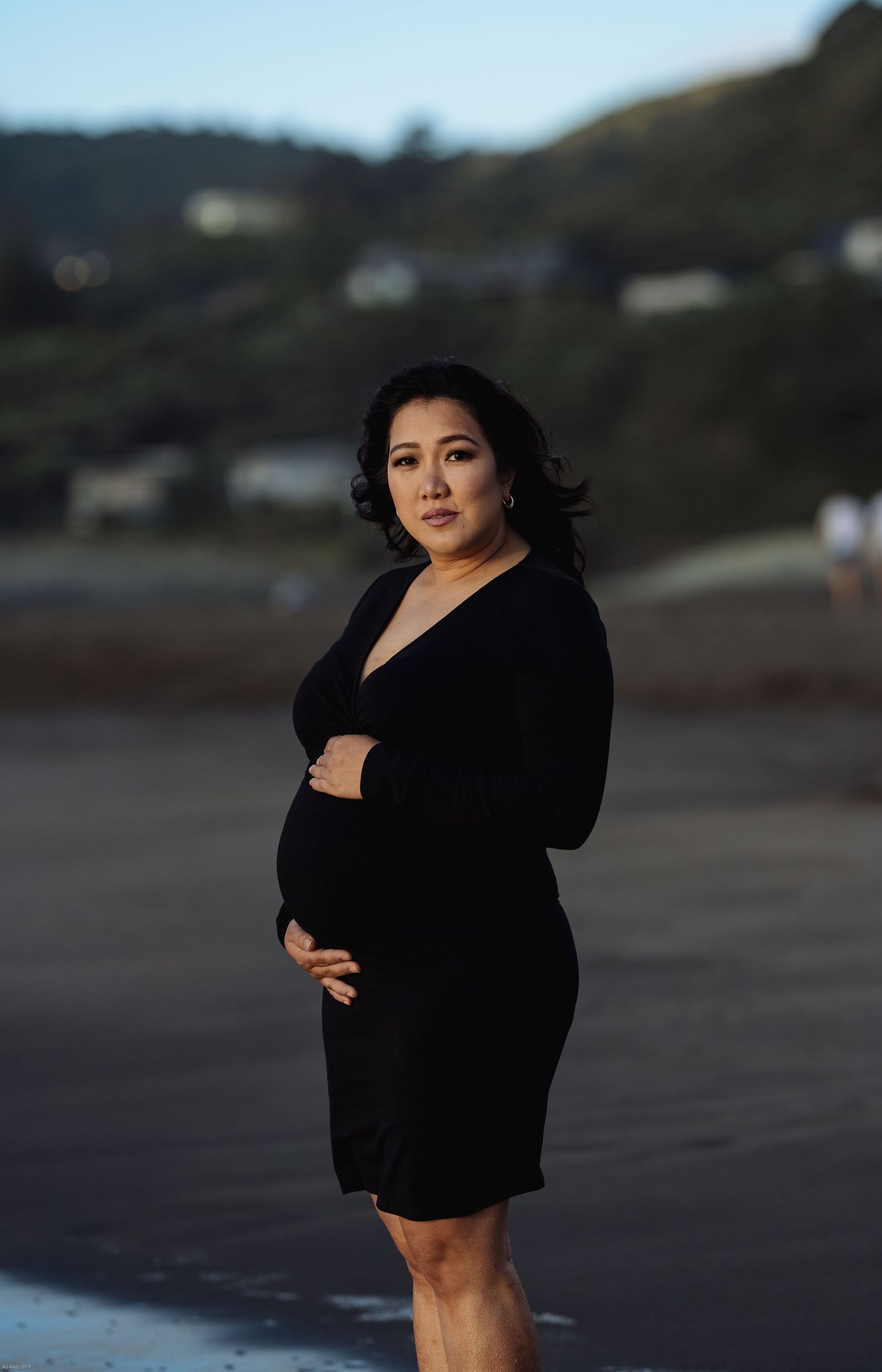 Person in black dress poses on moody beach with mountains in background during golden hour.