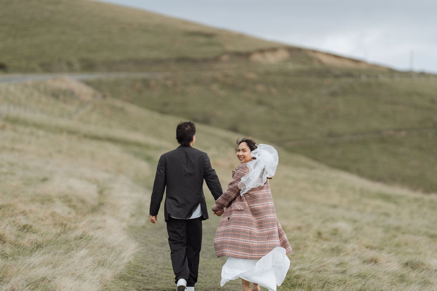 A couple walks together across rolling green hills in formal attire.