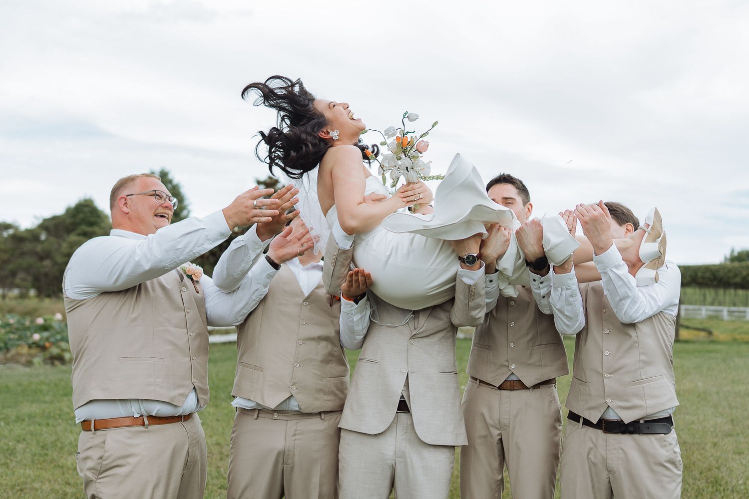 Wedding party members celebrate joyfully in light-colored attire outdoors.