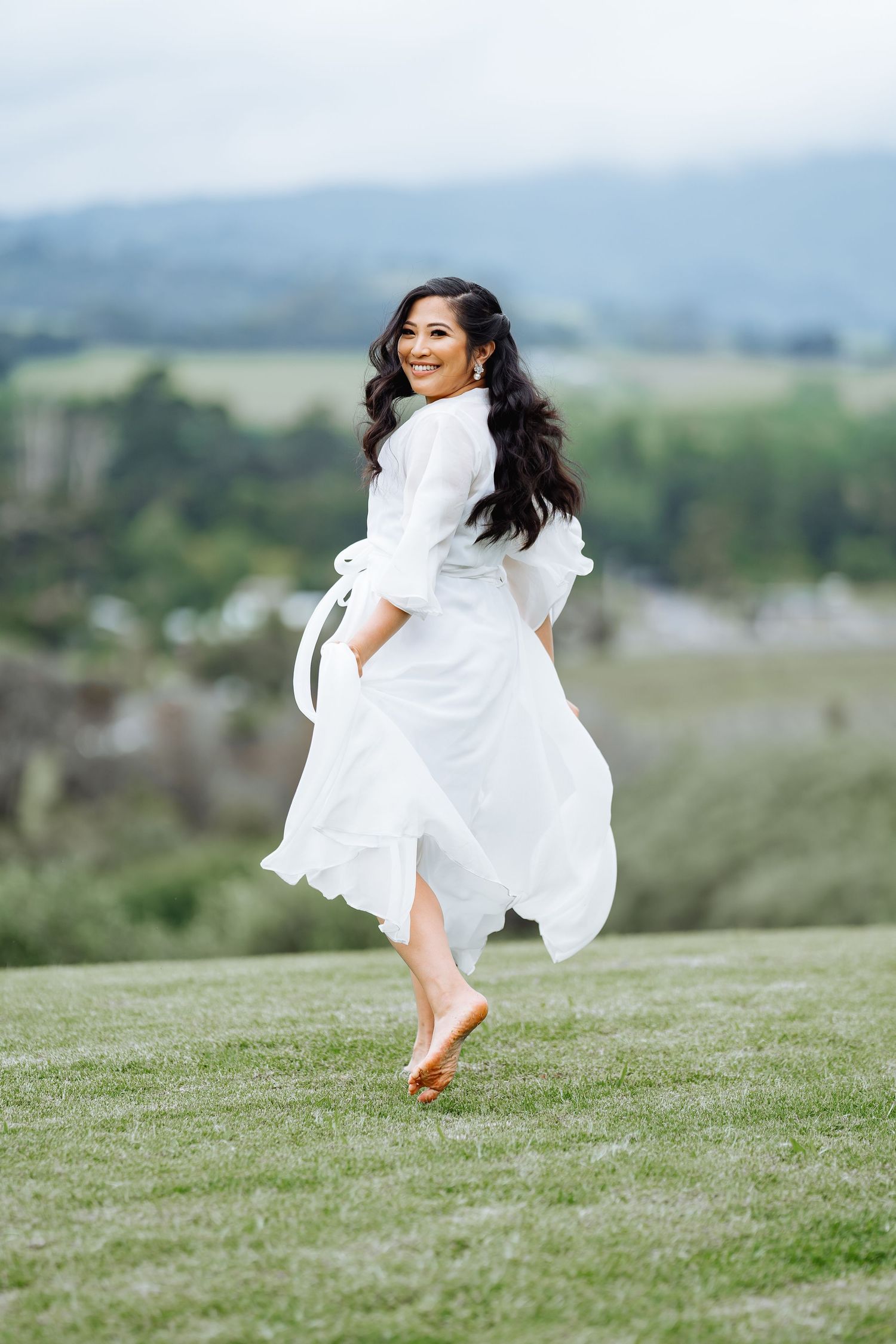 Bride in white dress twirling joyfully on a grassy hilltop with mountains in the background.