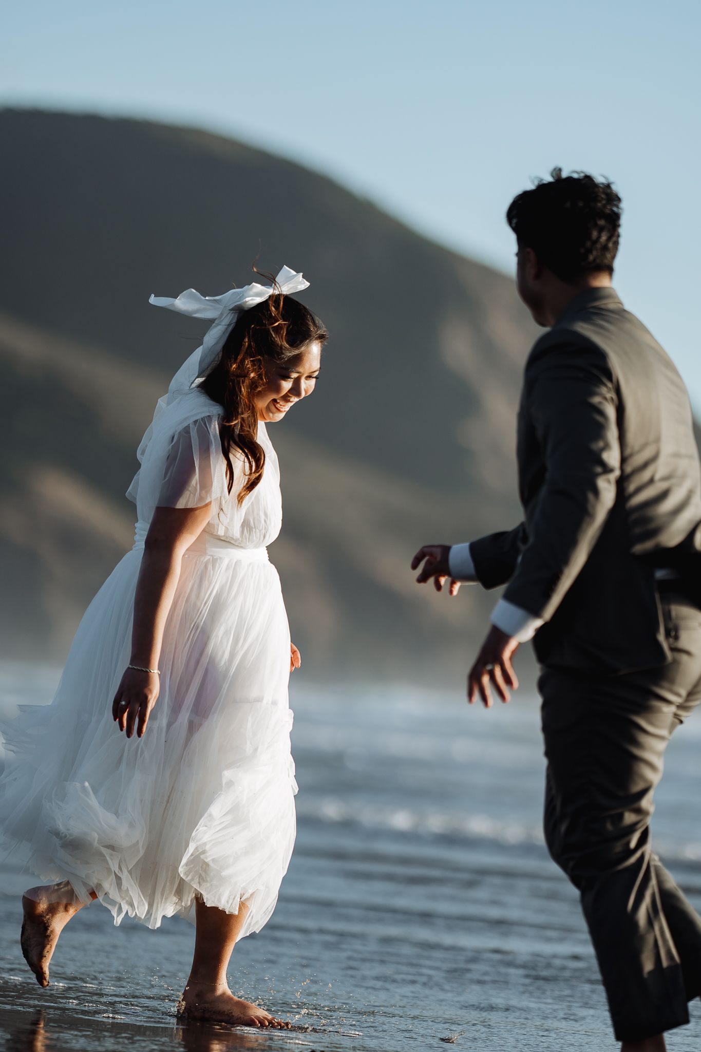A couple in wedding attire plays and dances on the beach near the water.