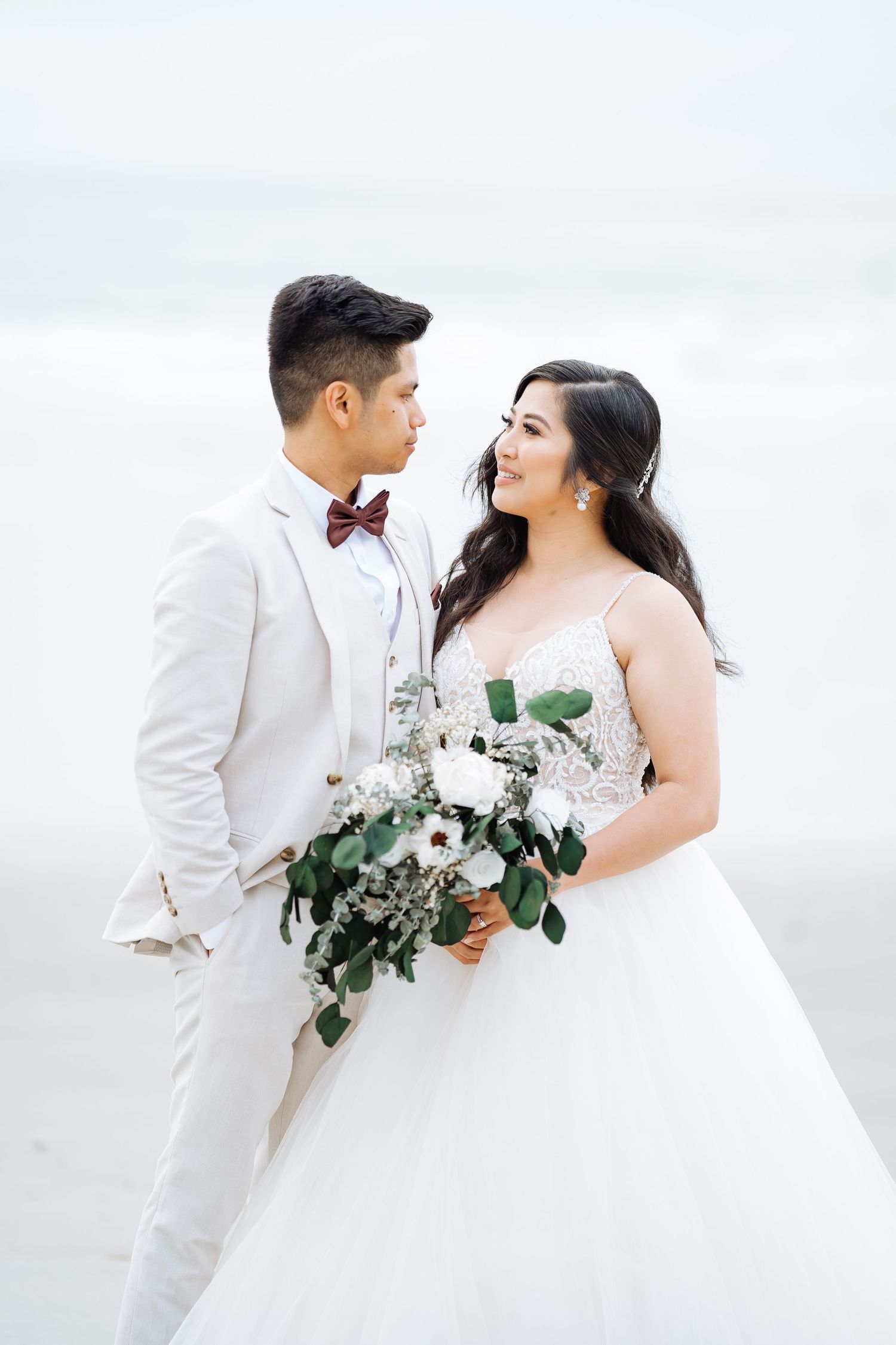 A couple in white wedding attire shares a romantic moment on a bright beach setting.