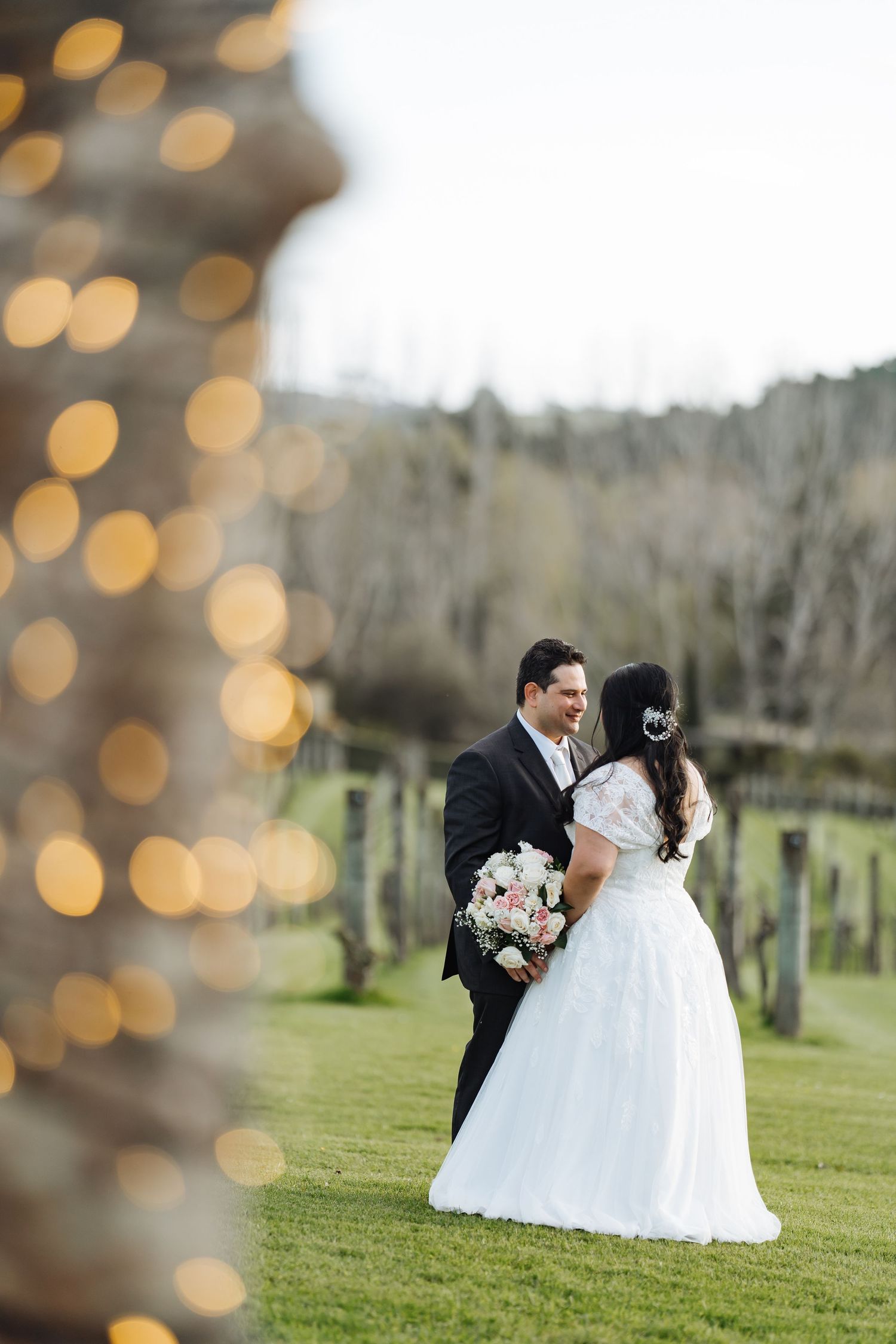 A wedding couple poses on a grassy field with bokeh lights in the foreground.