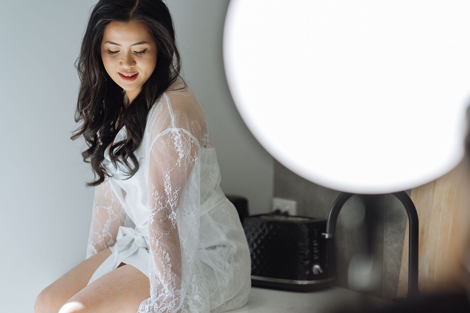 Bride in a long-sleeved lace wedding gown sitting by window light.