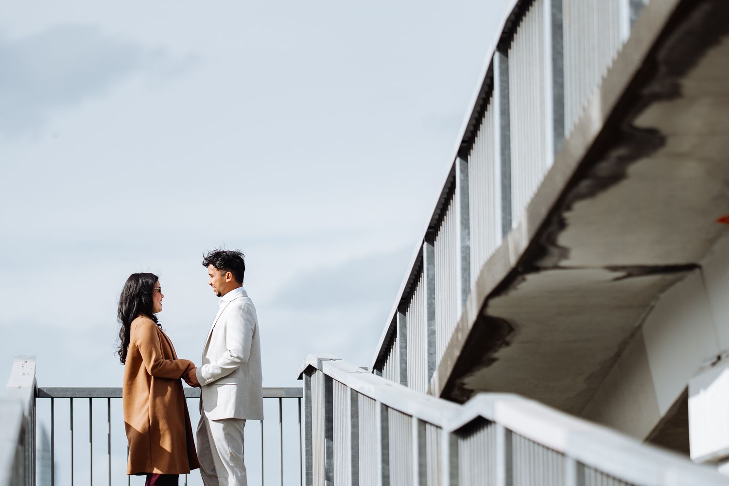 A couple stands on a white elevated walkway against a cloudy sky.