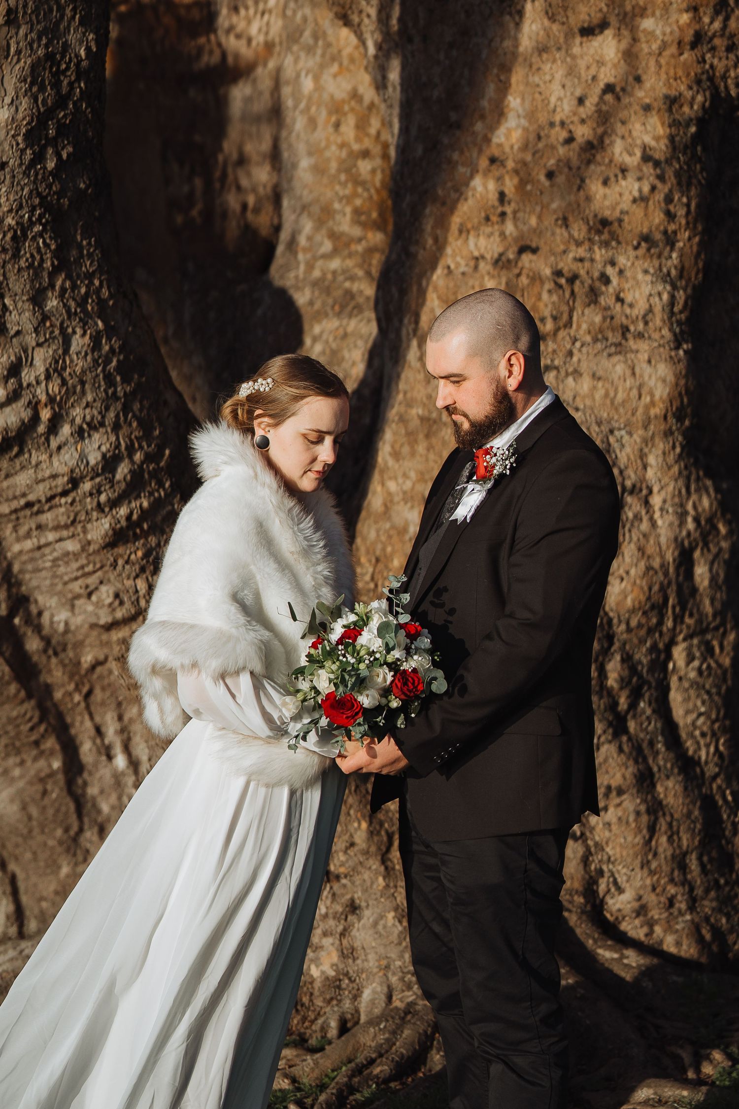 Romantic winter wedding couple poses against rustic tree backdrop with red floral bouquet.