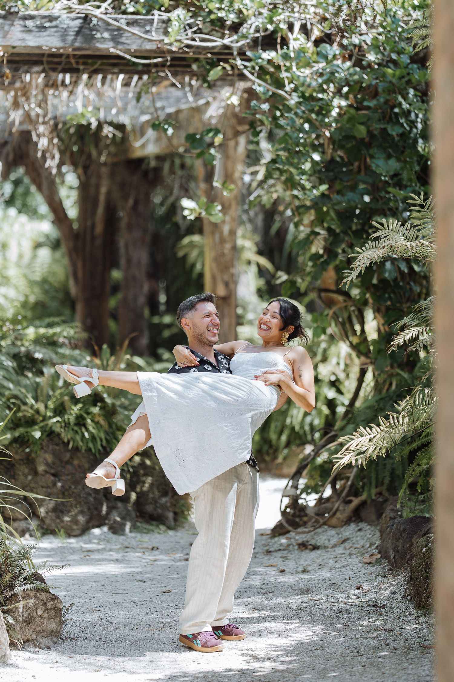 A couple shares a playful moment in a garden setting, with one person being lifted in a white dress.