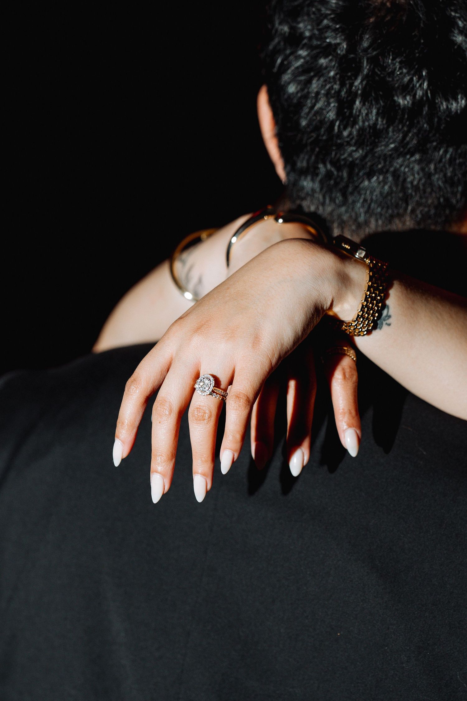 Close up of hands showing off an engagement ring and French manicure against a dark background.