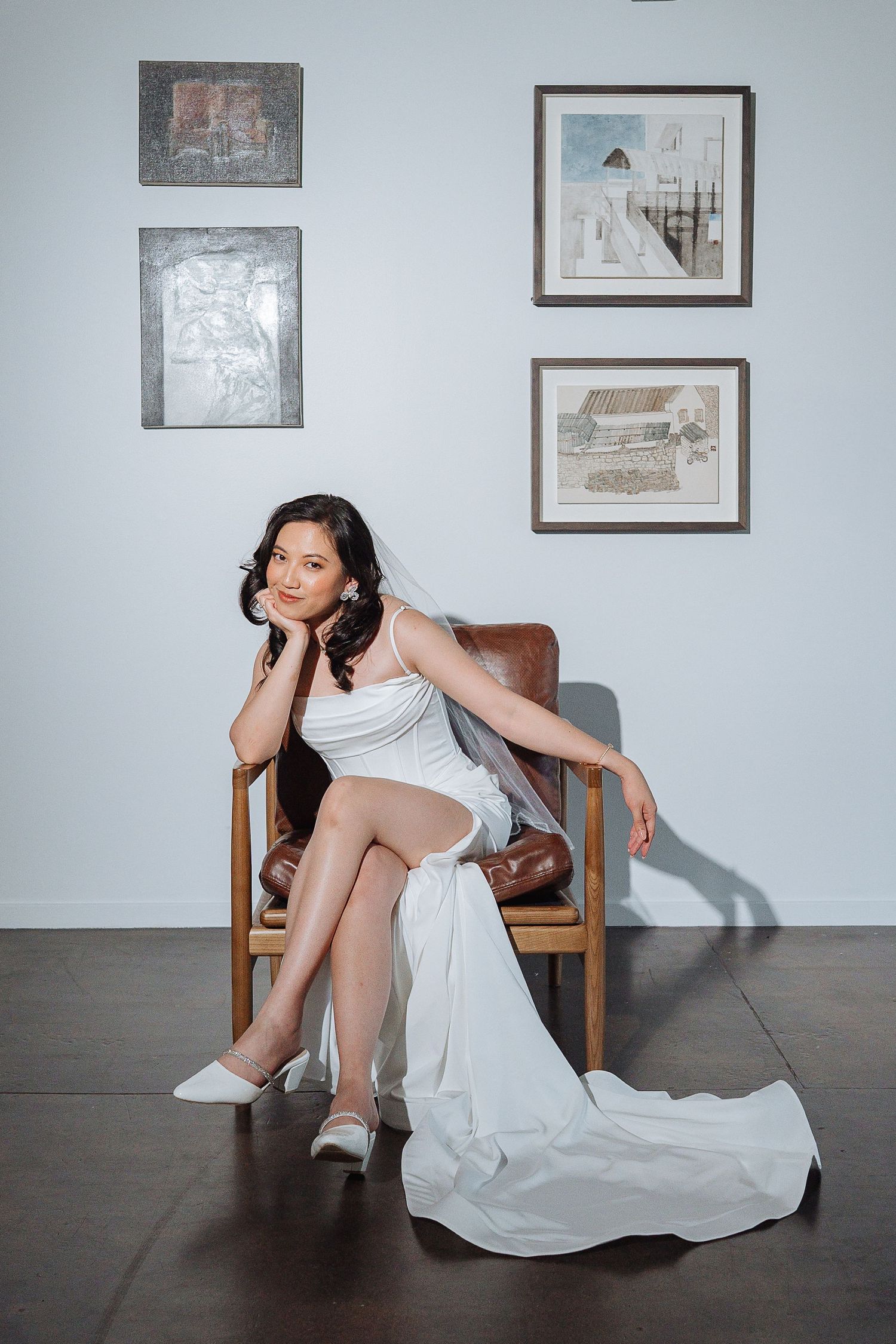 A figure in a flowing white dress poses elegantly in a chair against a wall with framed artwork.