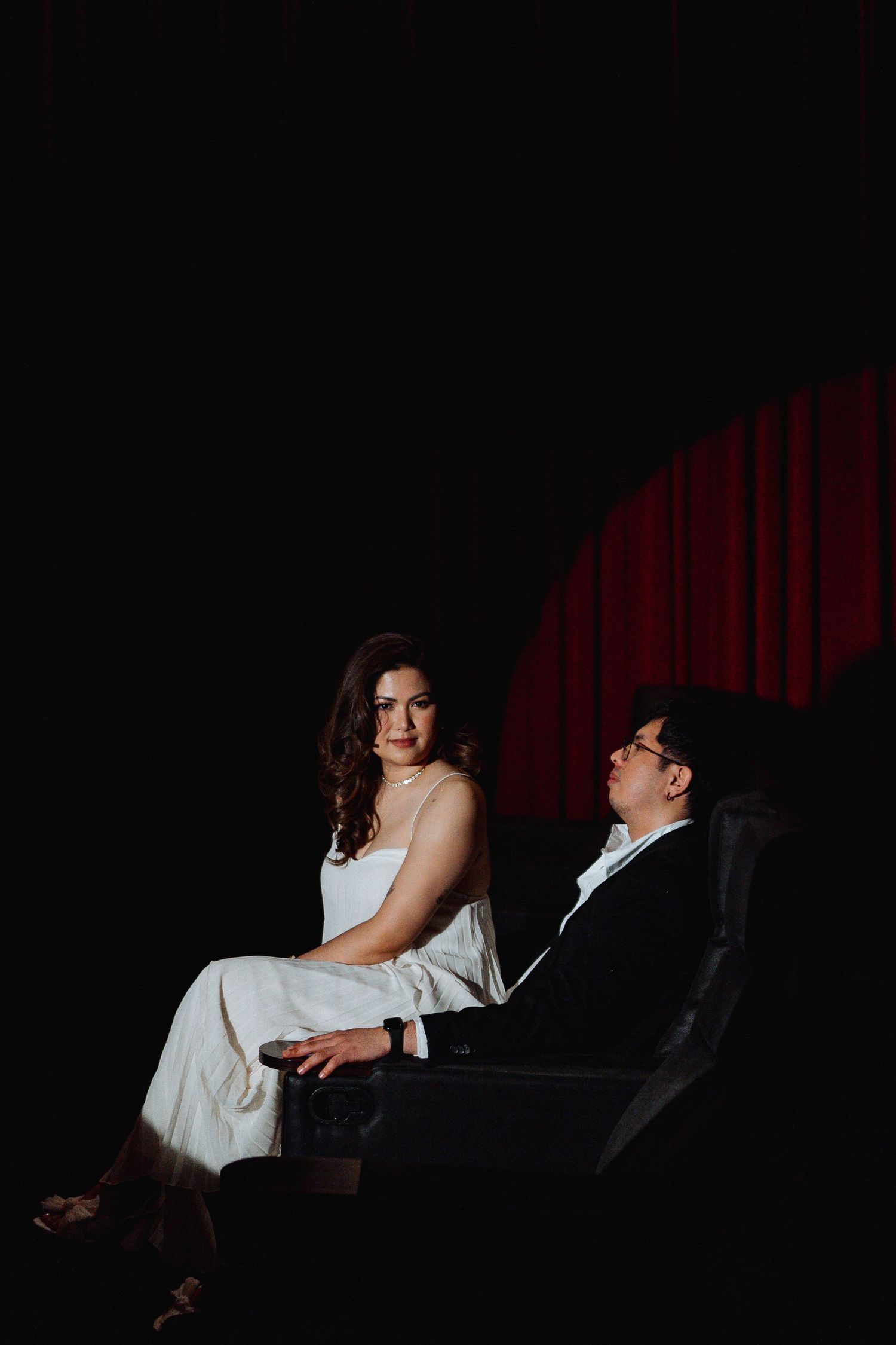 Couple in formal wear seated in dark theater with red curtain visible.