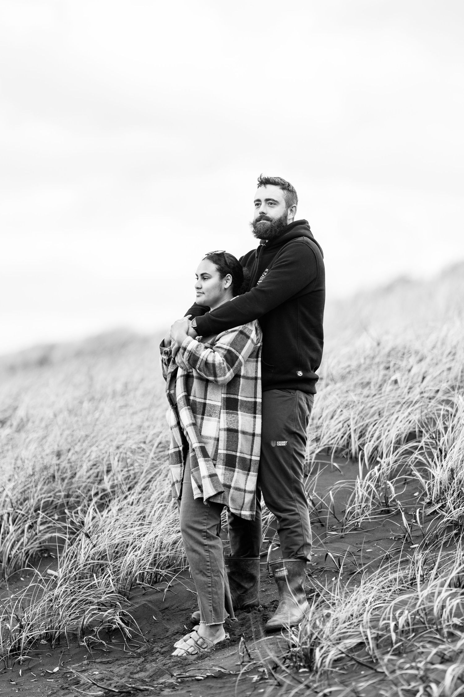 Black and white photo of a bearded man embracing a woman in a plaid shirt standing in tall grass outdoors.