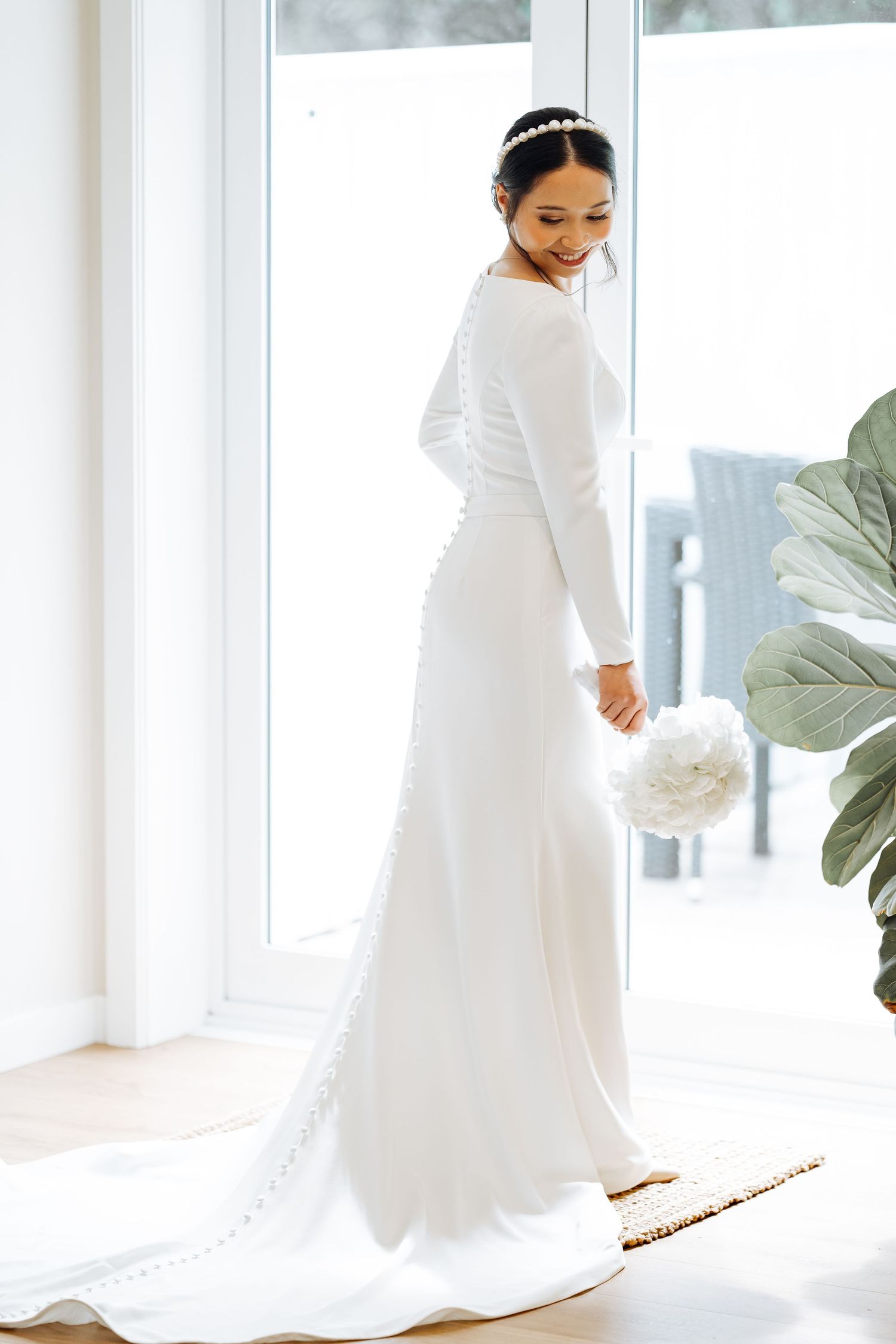 Bride in long-sleeved minimal wedding dress poses by window in natural light.