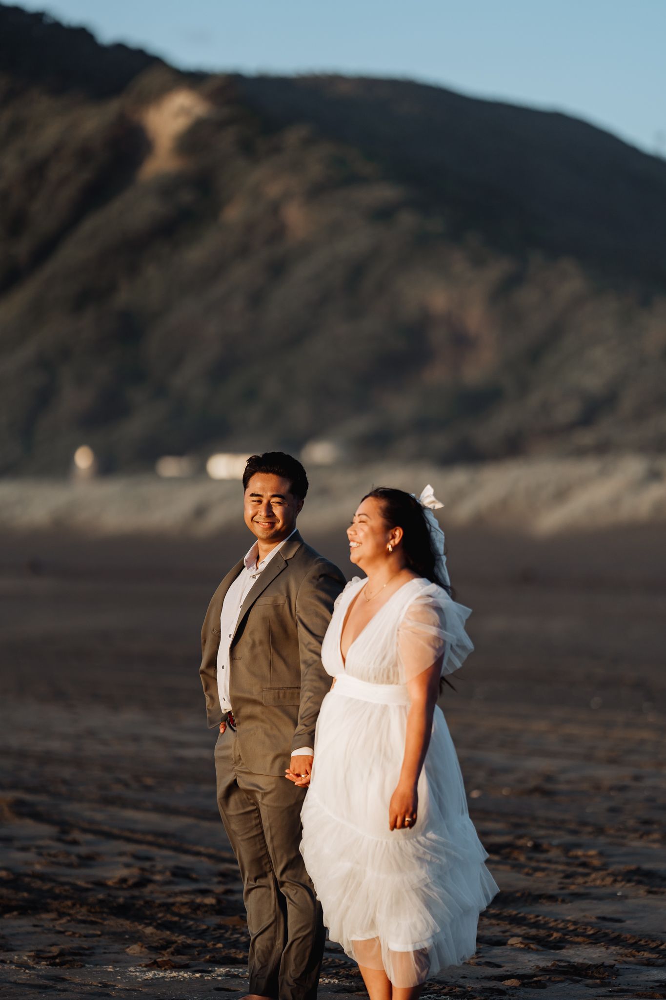 A couple in wedding attire laughs joyfully on a beach with mountains in the background.