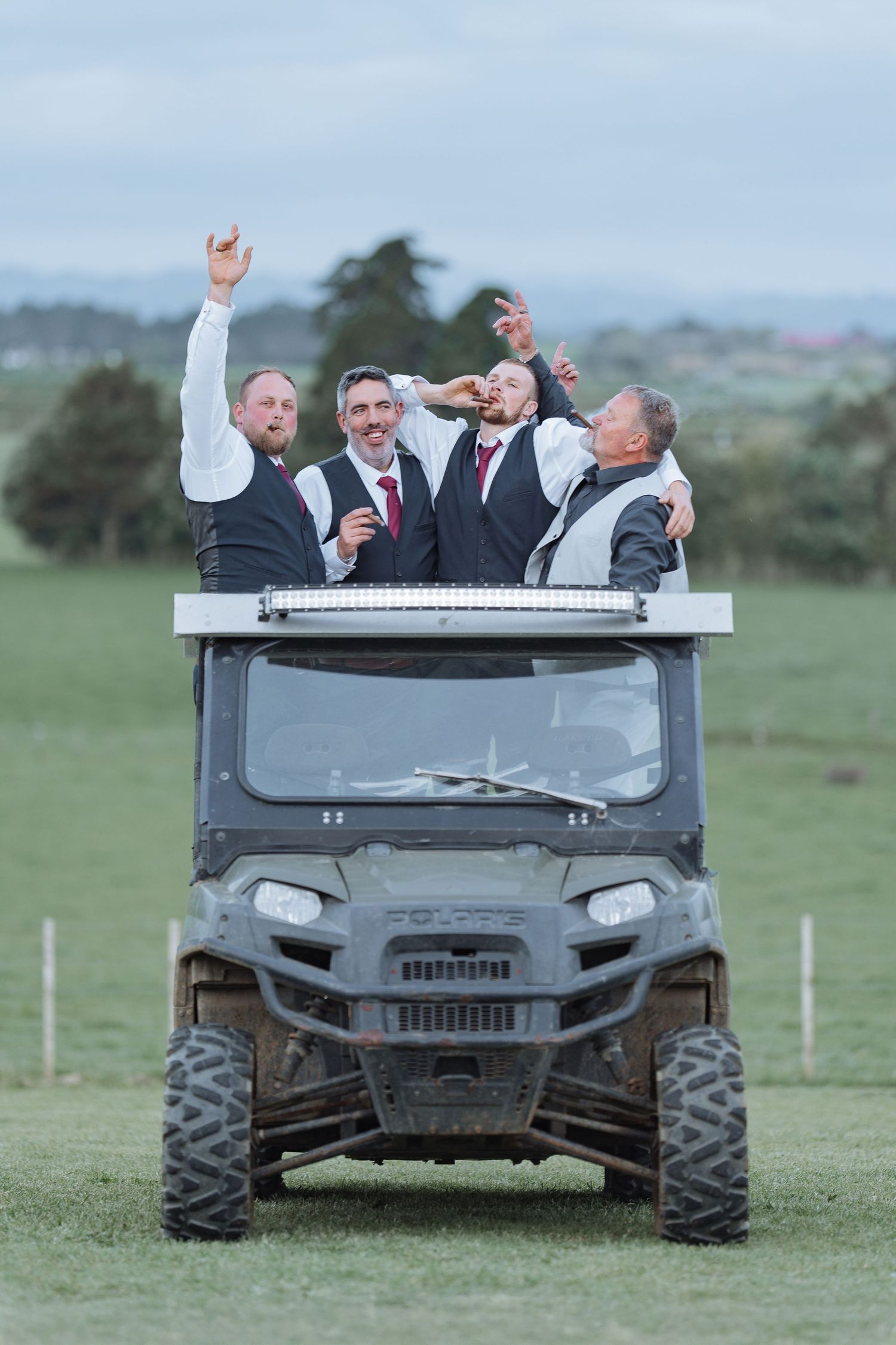 Groomsmen celebrating on a utility vehicle in an open field during wedding festivities.
