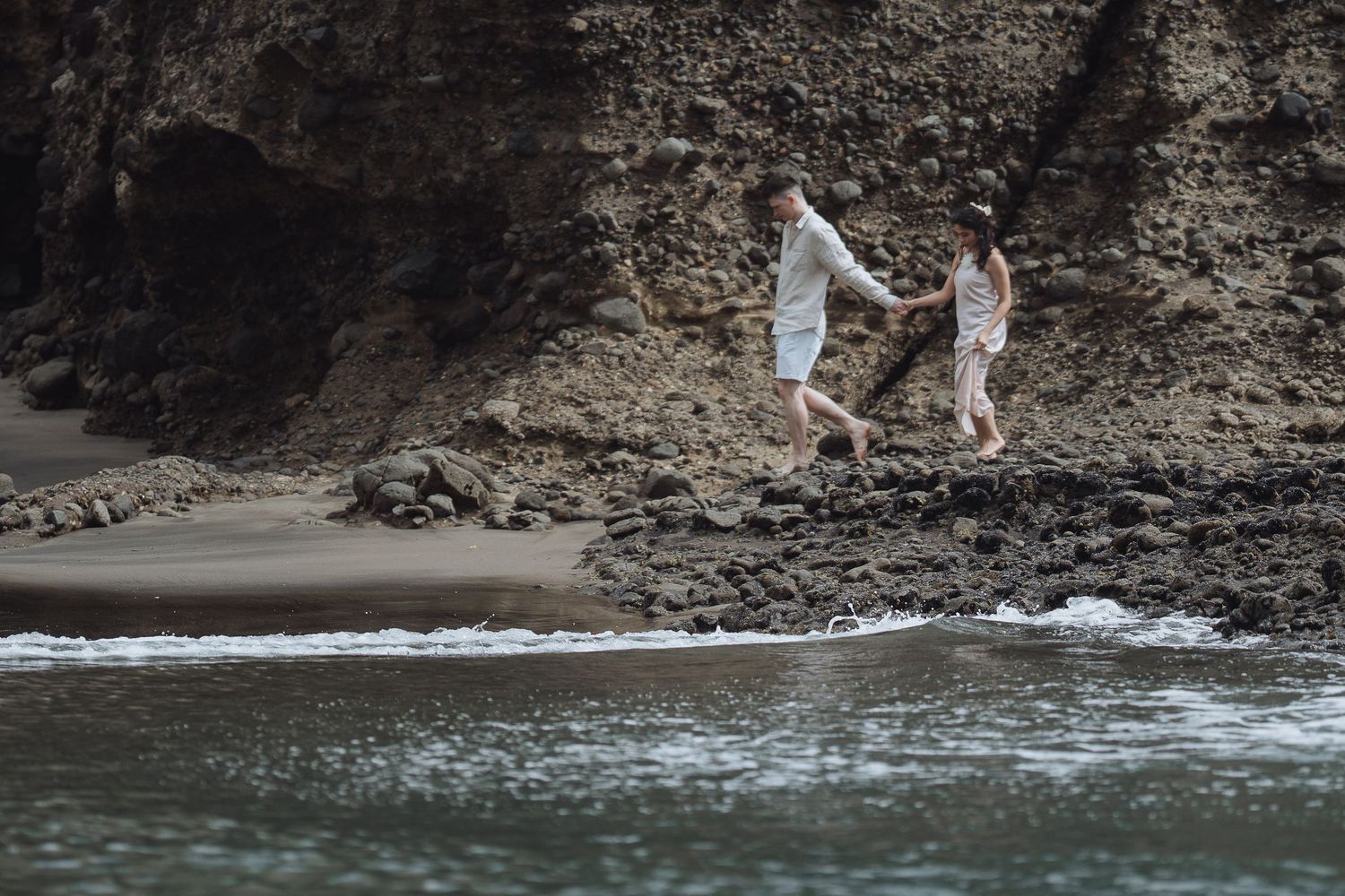Couple walking along a rocky coastline with crashing waves and dramatic cliffs in the background.