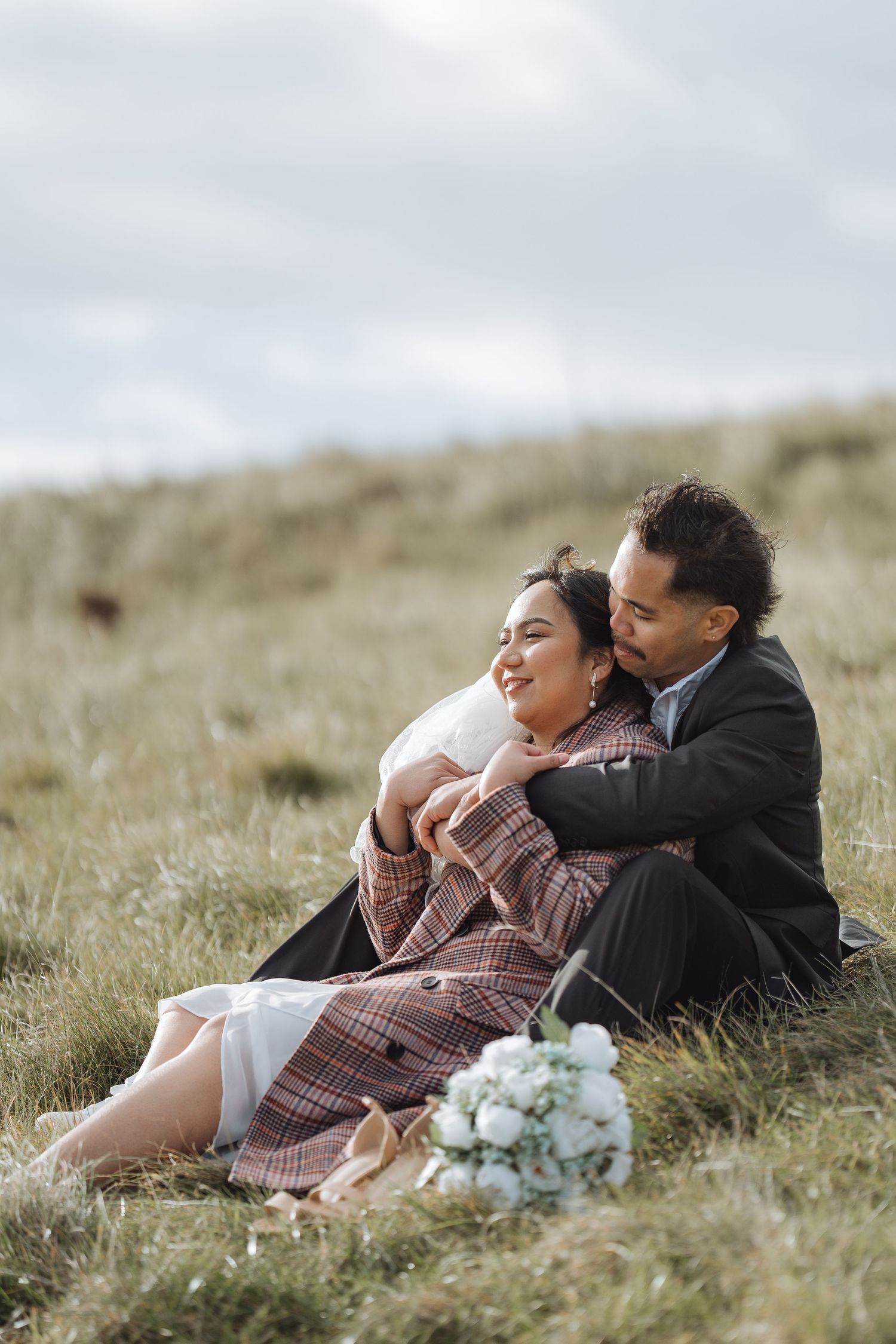 A couple relaxes together on a grassy hillside with white flowers nearby.