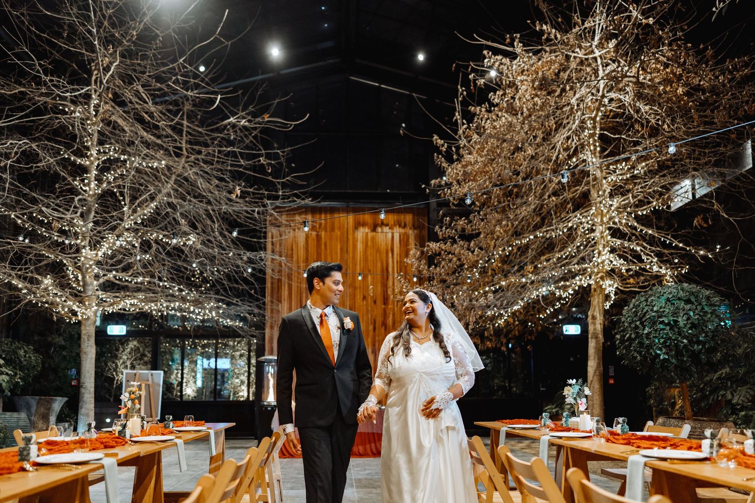 Wedding reception tables decorated with candles and floral arrangements under illuminated trees at night.