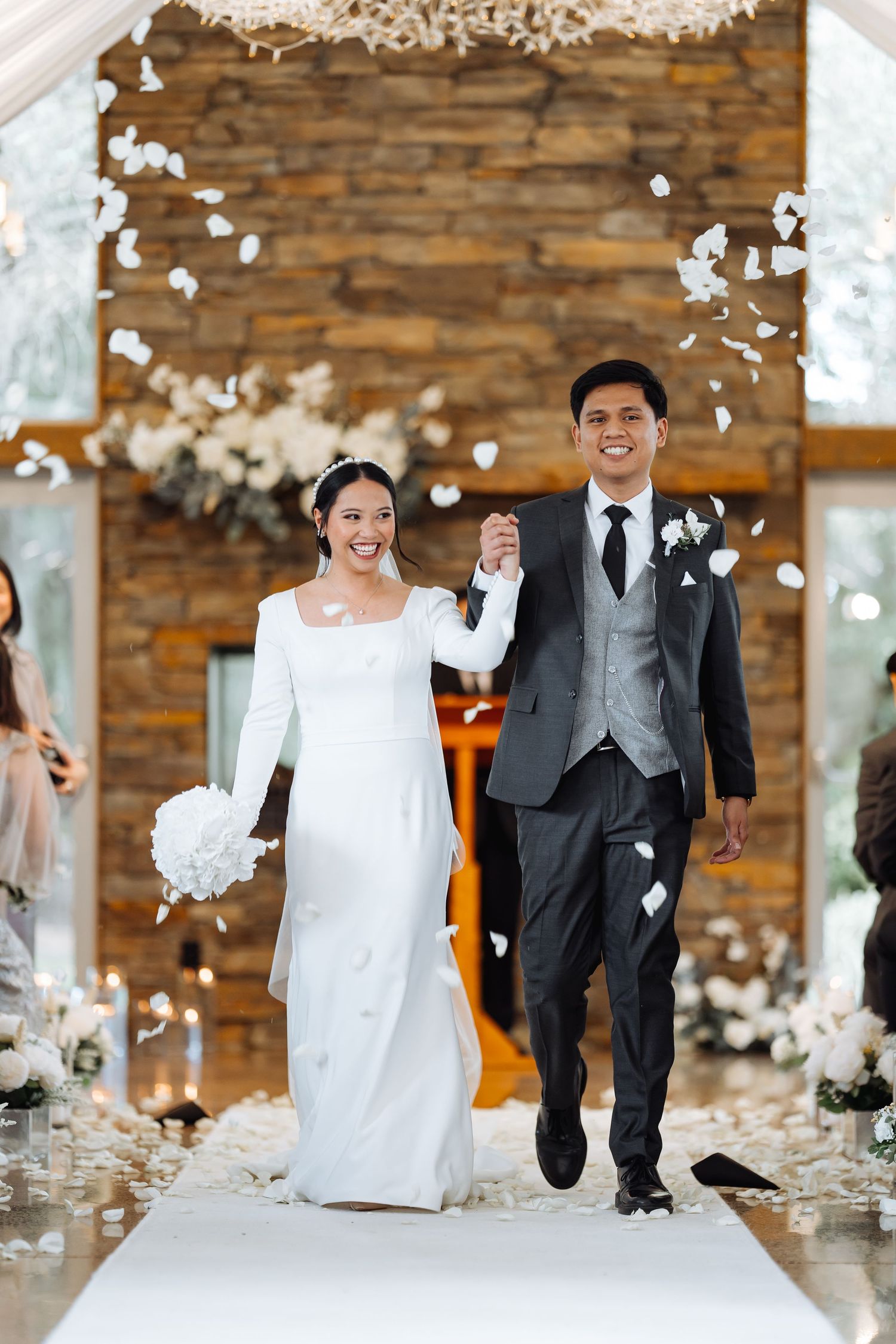 Newlyweds walking down aisle amid falling flower petals against rustic wooden backdrop.
