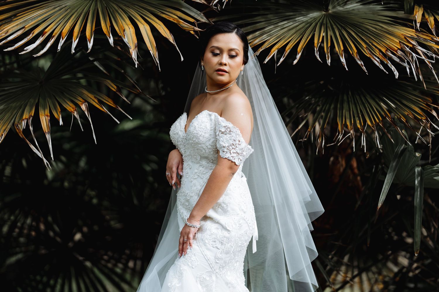 Elegant bride in a lace off-shoulder wedding dress poses against tropical palm fronds in a garden setting.