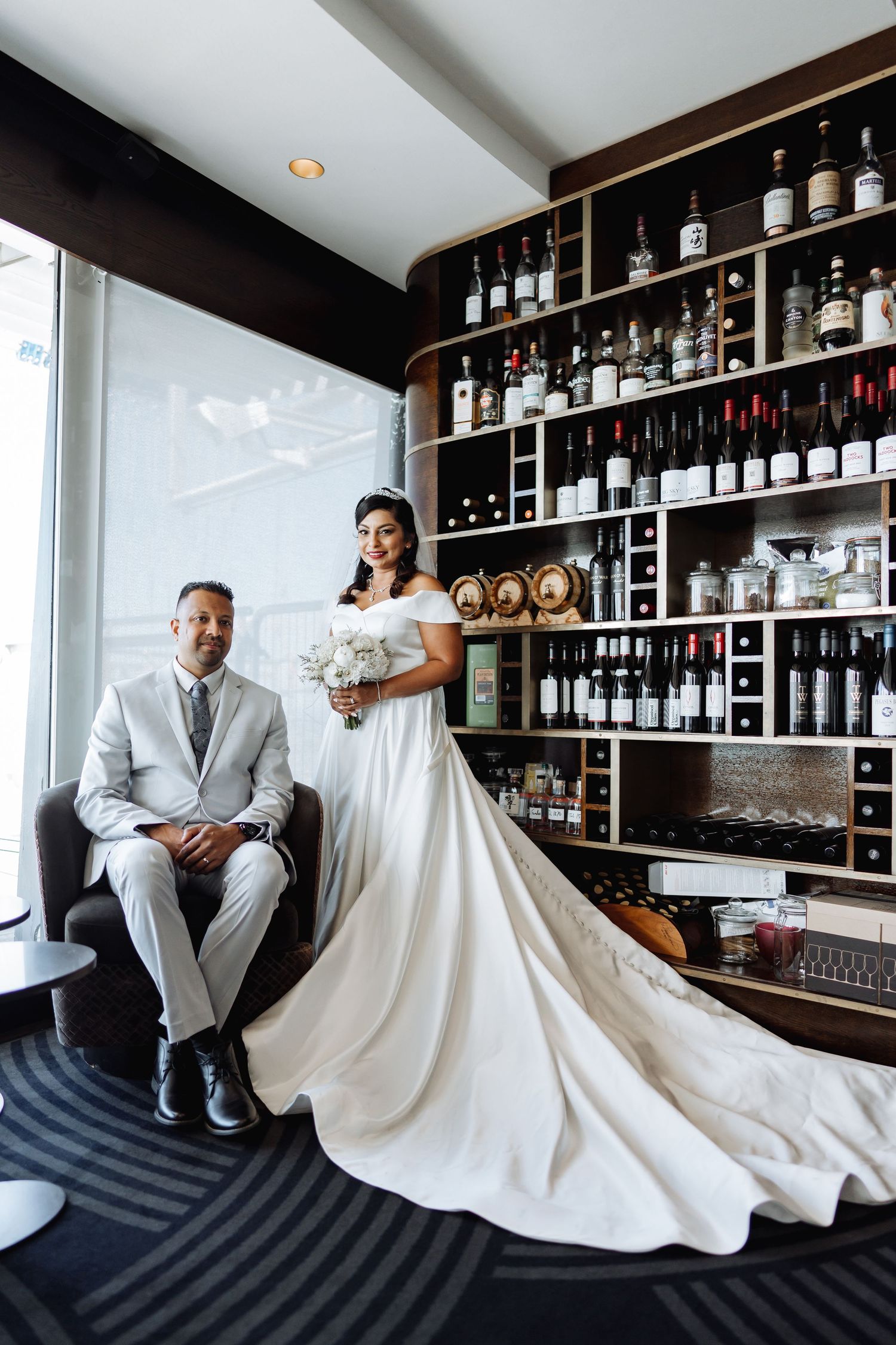 Elegant wedding portrait in upscale bar with dramatic flowing white dress and wall of bottles.