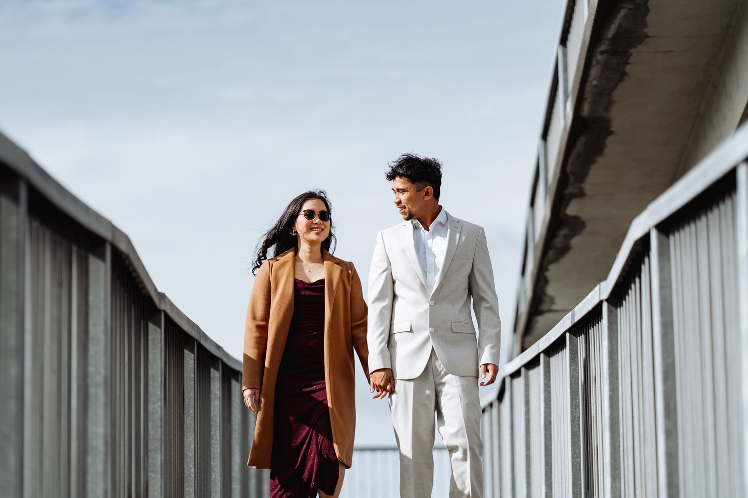 Romantic couple walking on a modern white railing pathway against a cloudy sky.