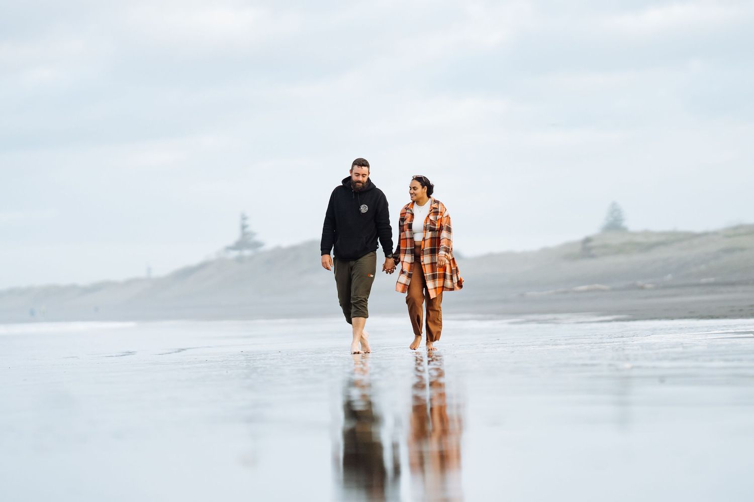 Couple walking barefoot on a wet reflective beach on a cloudy day, wearing casual autumn outfits.