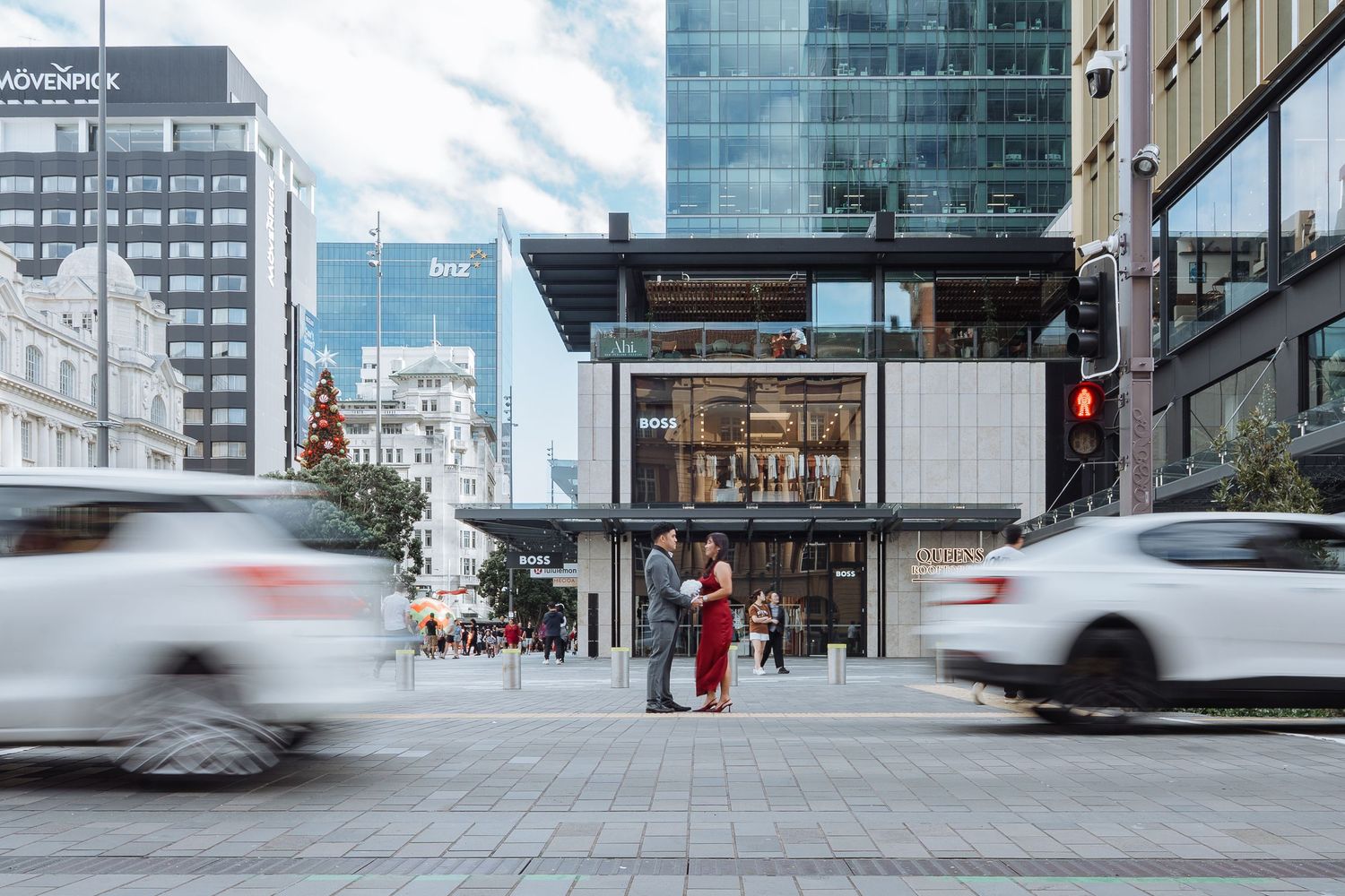 Urban street scene with motion blur of passing vehicles and pedestrians against modern city buildings.