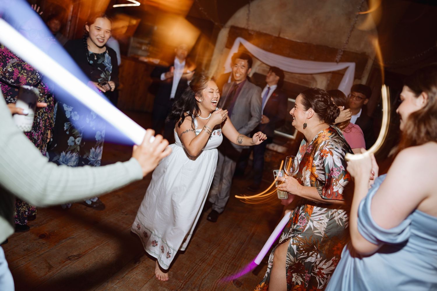 Party guests dance and celebrate under vibrant purple lighting at an indoor reception venue.