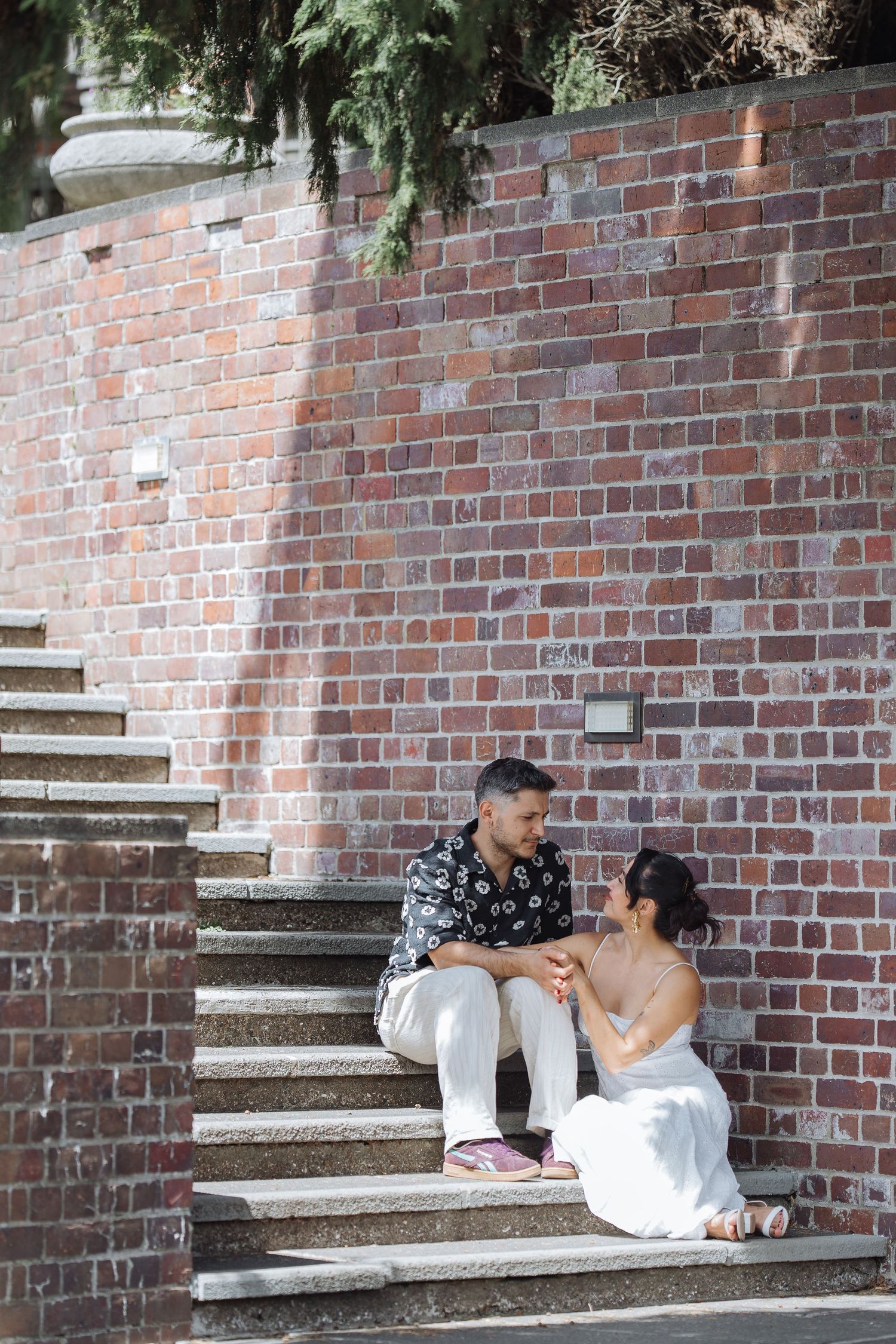 A couple poses playfully on brick steps surrounded by greenery and architecture.