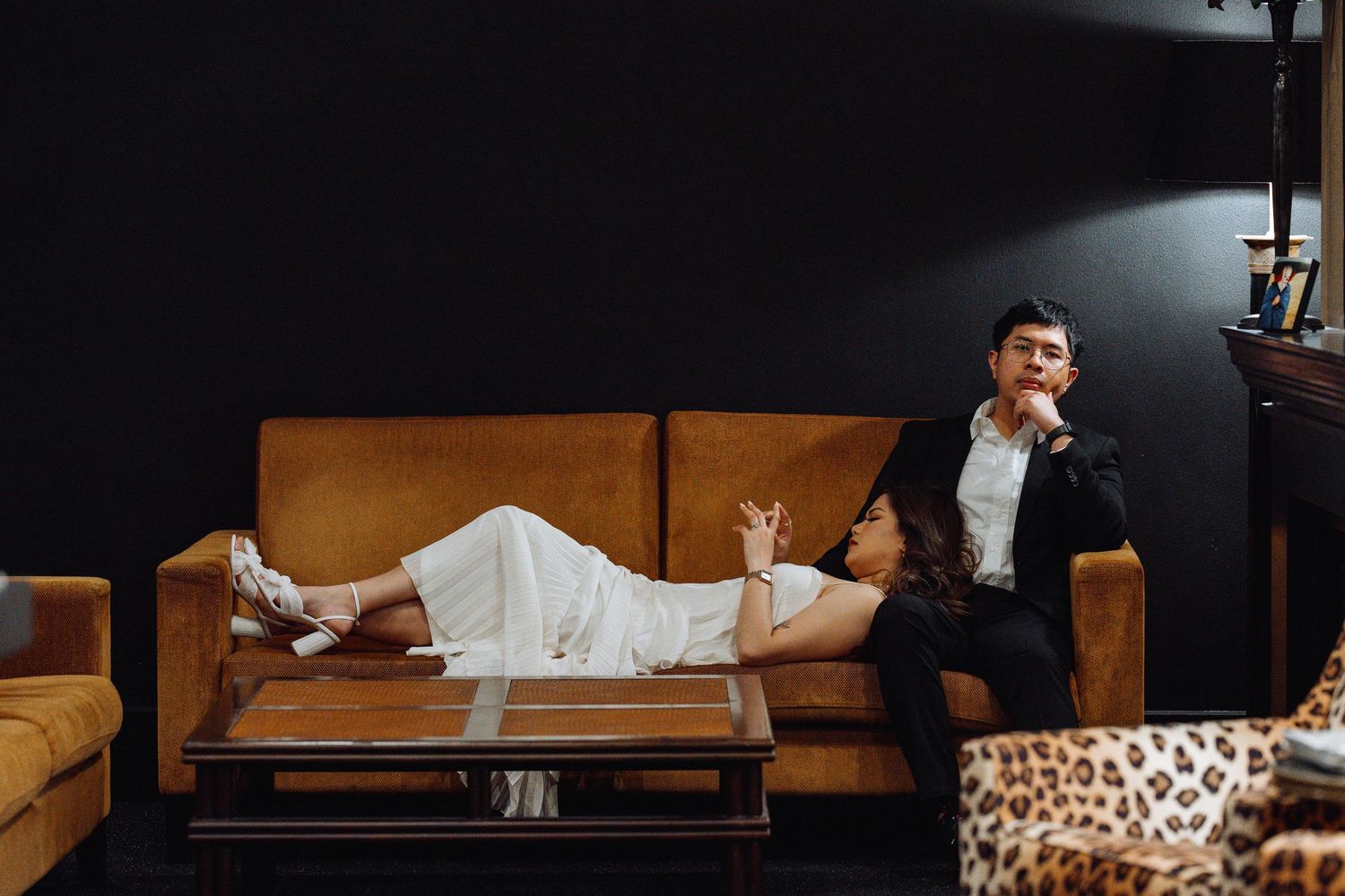 A couple poses dramatically on a brown leather couch in a moody studio setting.