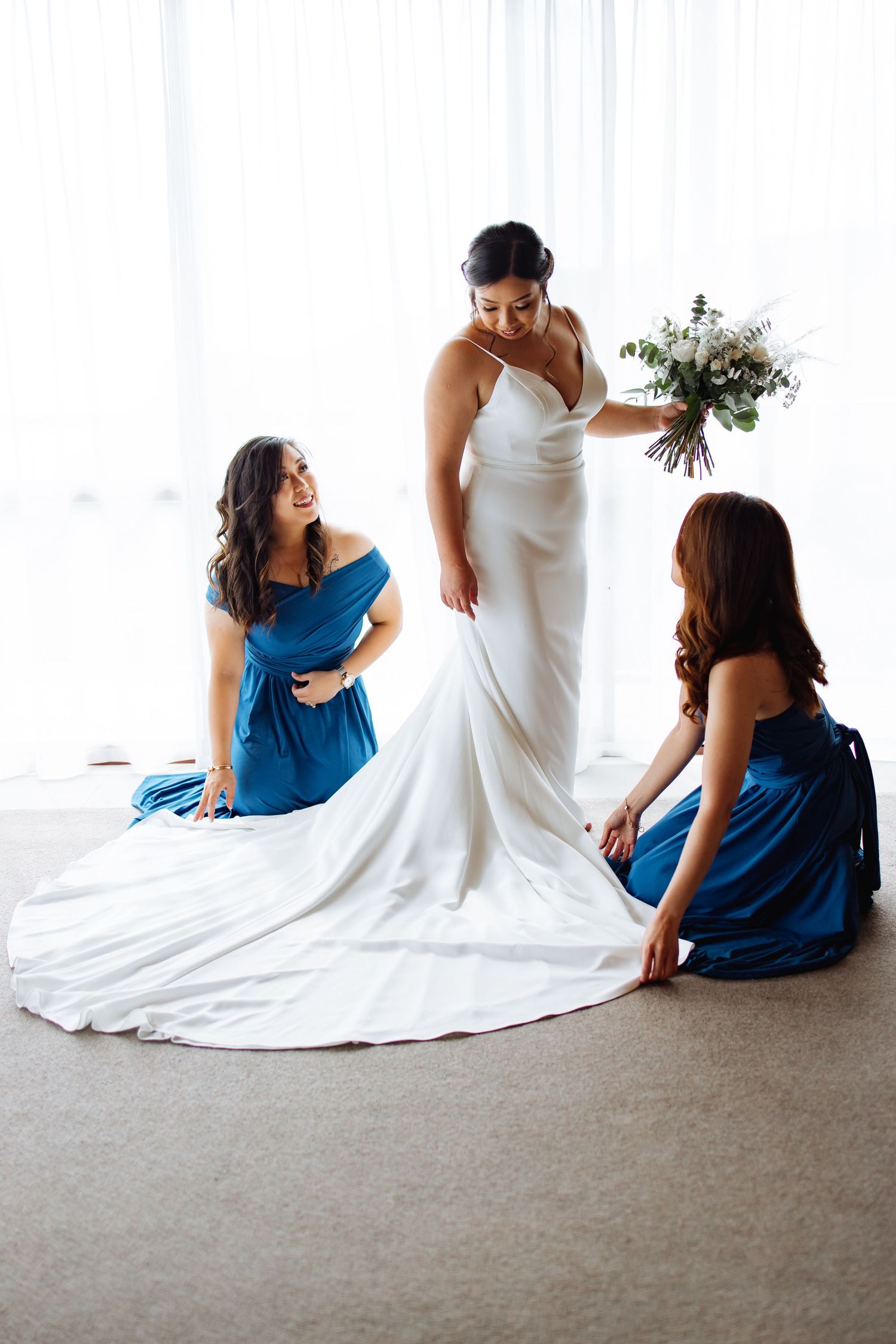 Bridesmaids in blue dresses help adjust bride's white wedding gown on carpeted floor.