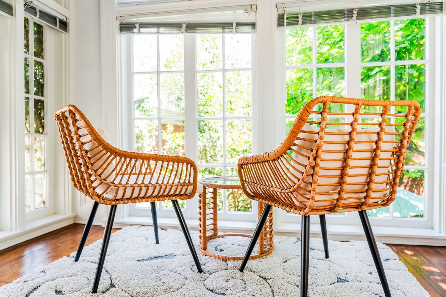 Two woven rattan chairs with black metal legs sit by bright windows in a sunlit room with white trim and hardwood floors.
