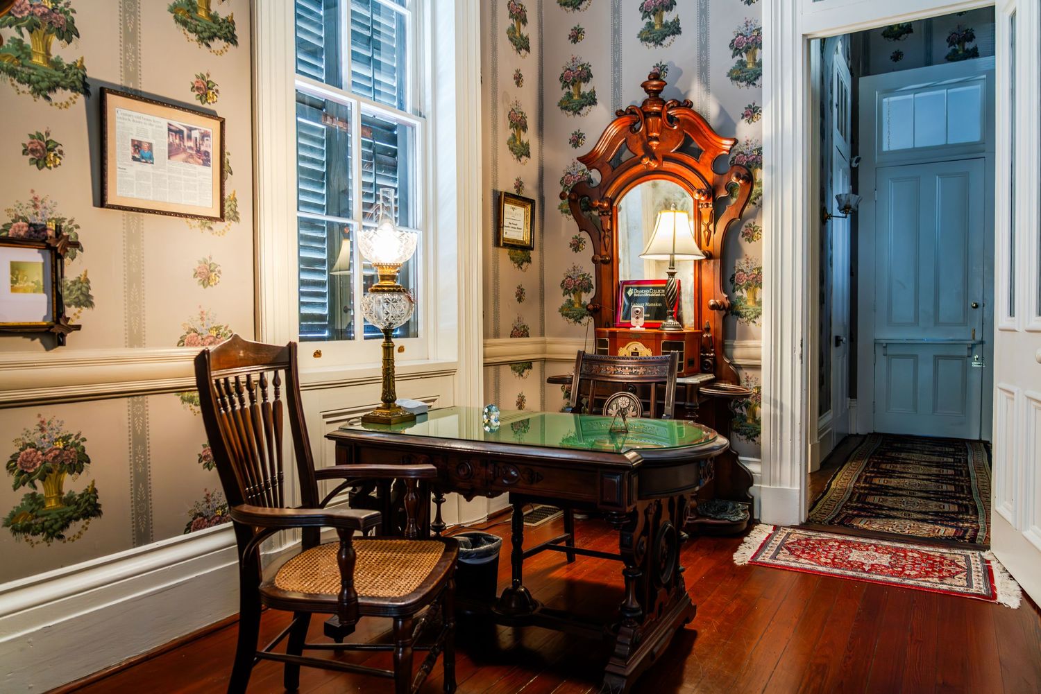 Vintage desk with ornate mirror and wooden chair sits against floral wallpapered walls in historic room with hardwood floors.
